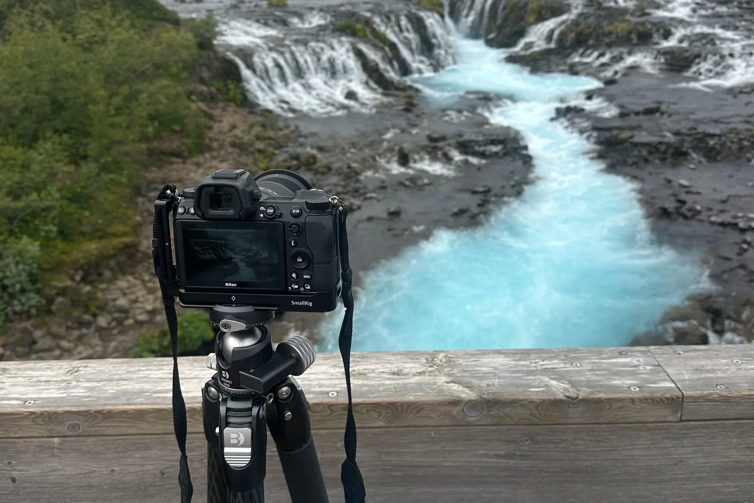 A DSLR camera on a tripod facing a river with waterfalls and greenery in the background.