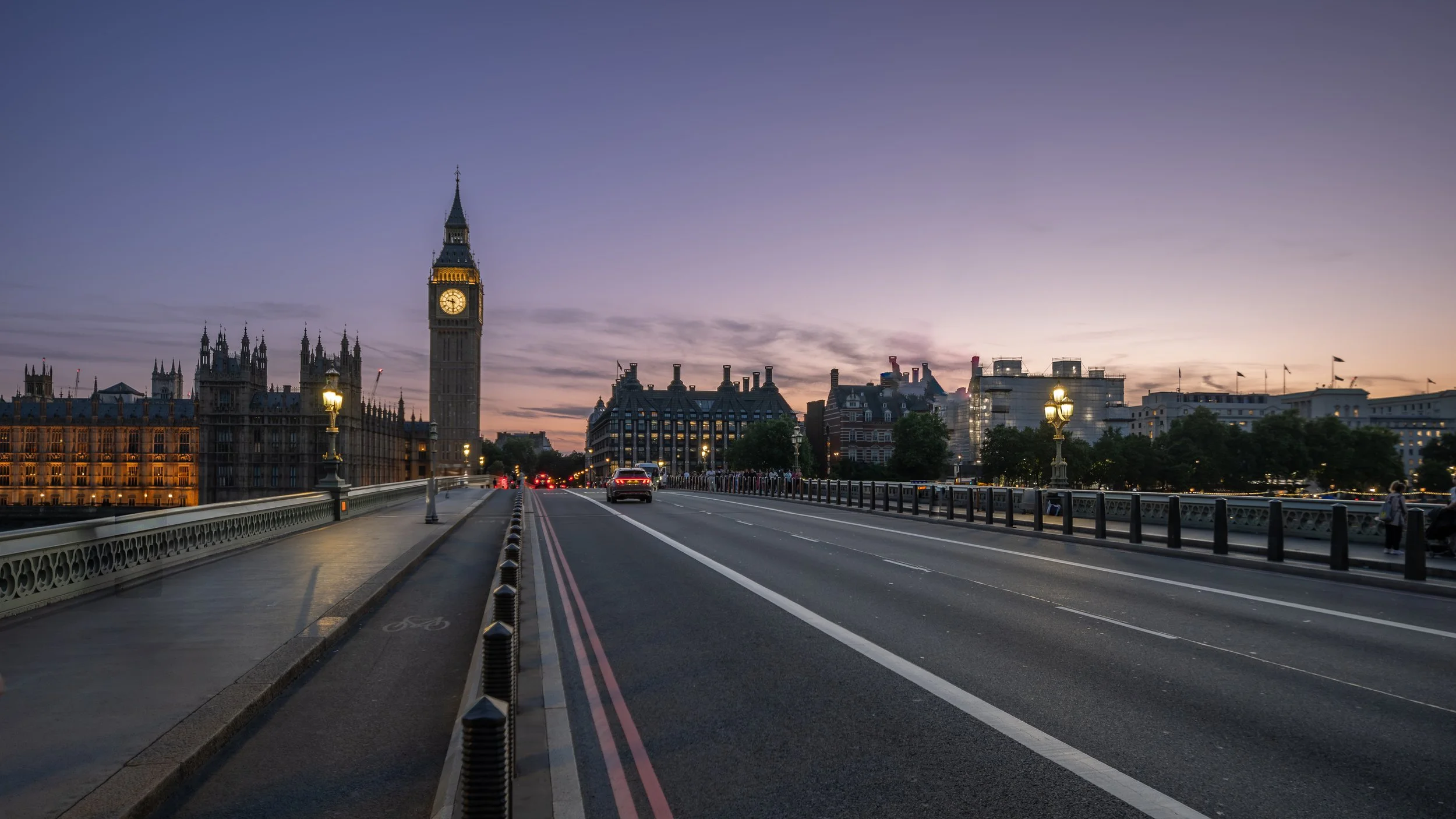 Westminster Bridge at dusk with the iconic clock tower on the left, streetlights glowing, and a serene, lilac sky enhancing the historic architecture.
