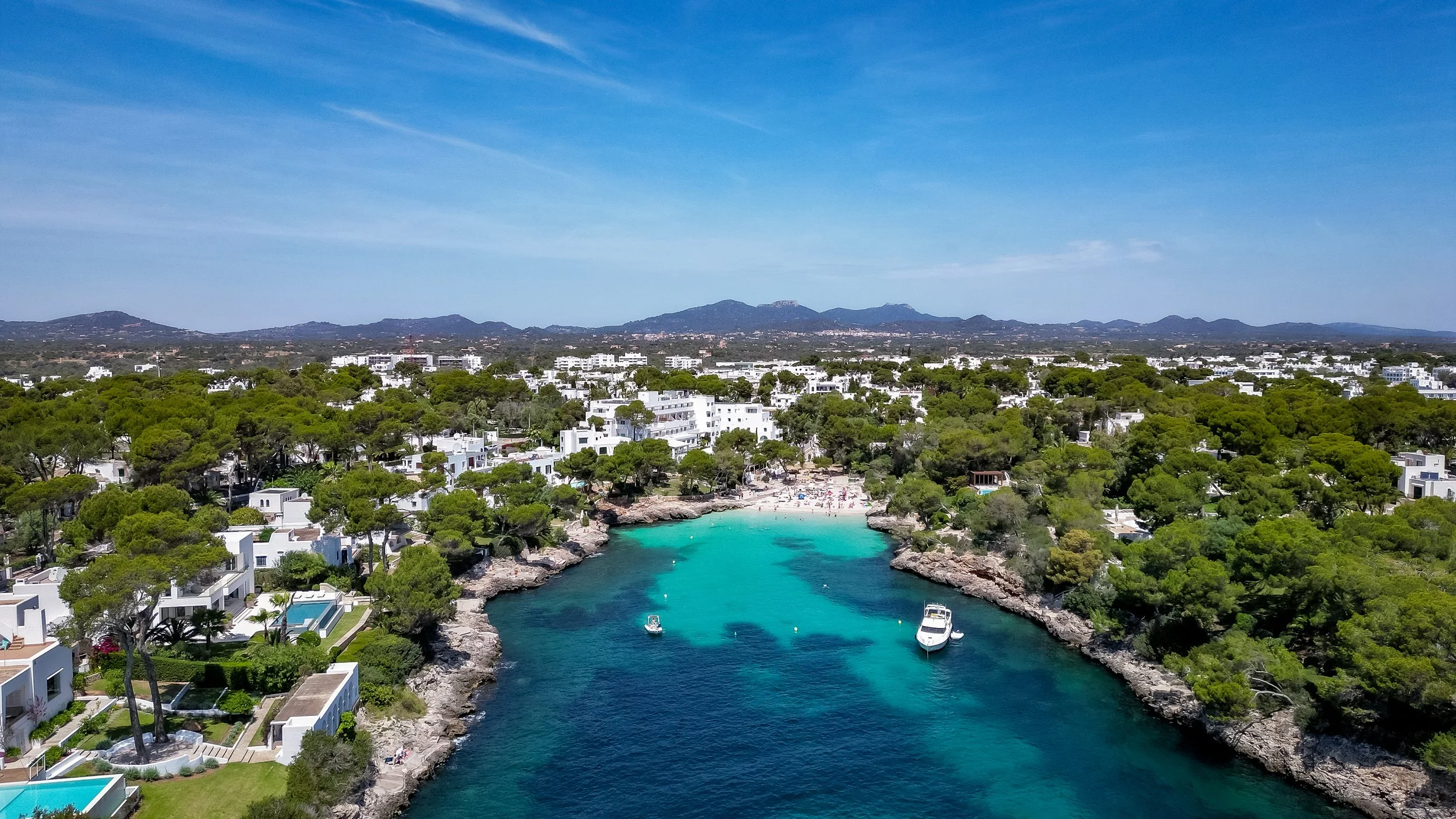 Aerial view of a coastal town with white buildings, green trees, blue water, and boats in a small bay on a sunny day.