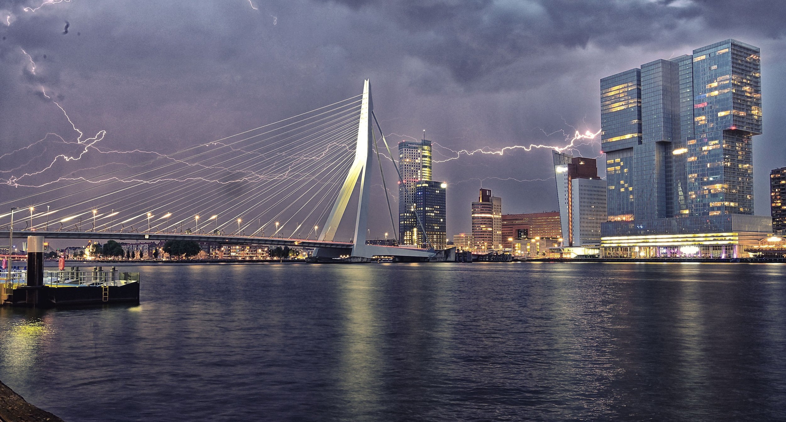 Nighttime cityscape of Rotterdam with lightning in the stormy sky, modern skyscrapers, and the Erasmus Bridge over the water.