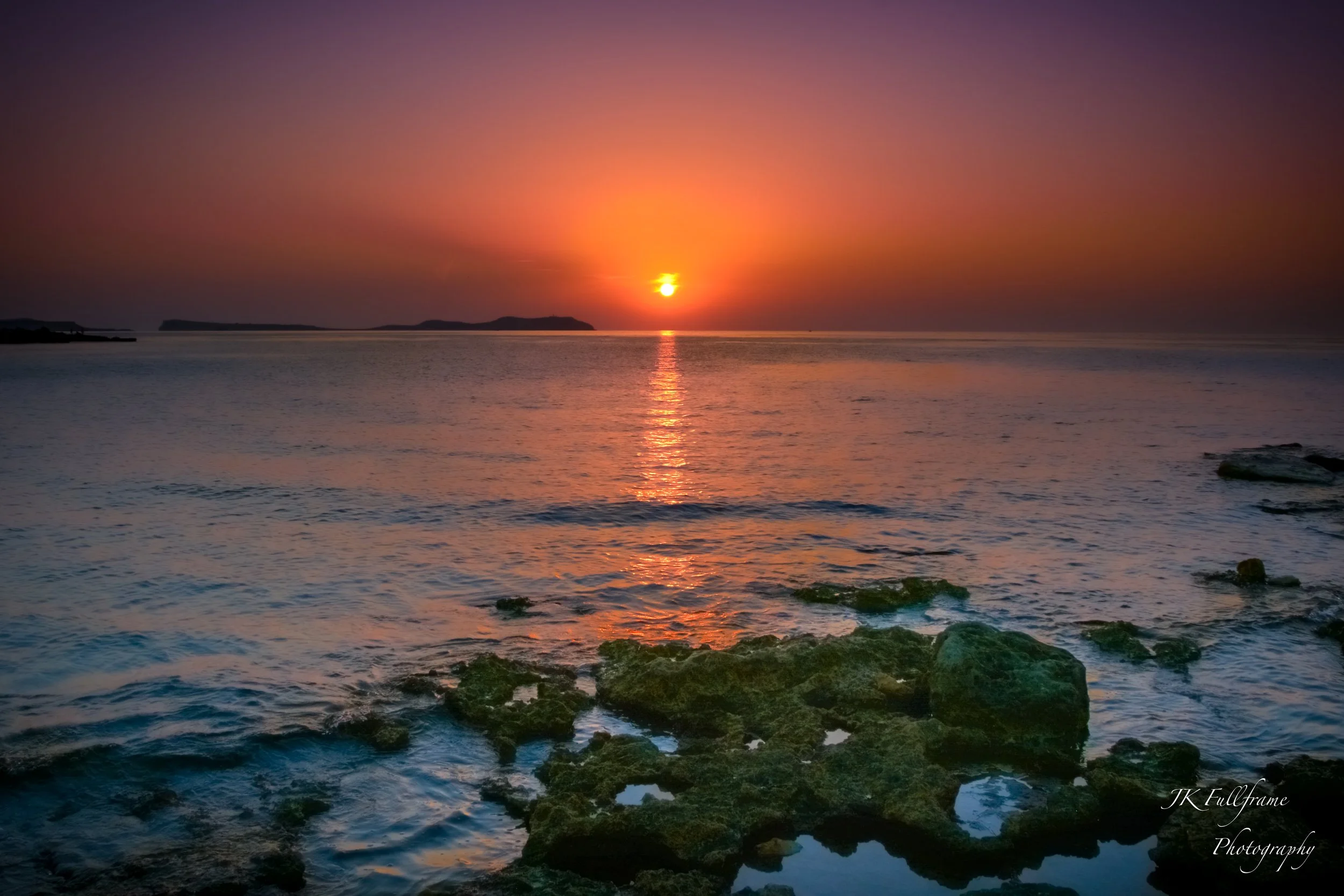 Sunset over the ocean with colorful sky, rocky shoreline in foreground, calm water reflecting the sunset.