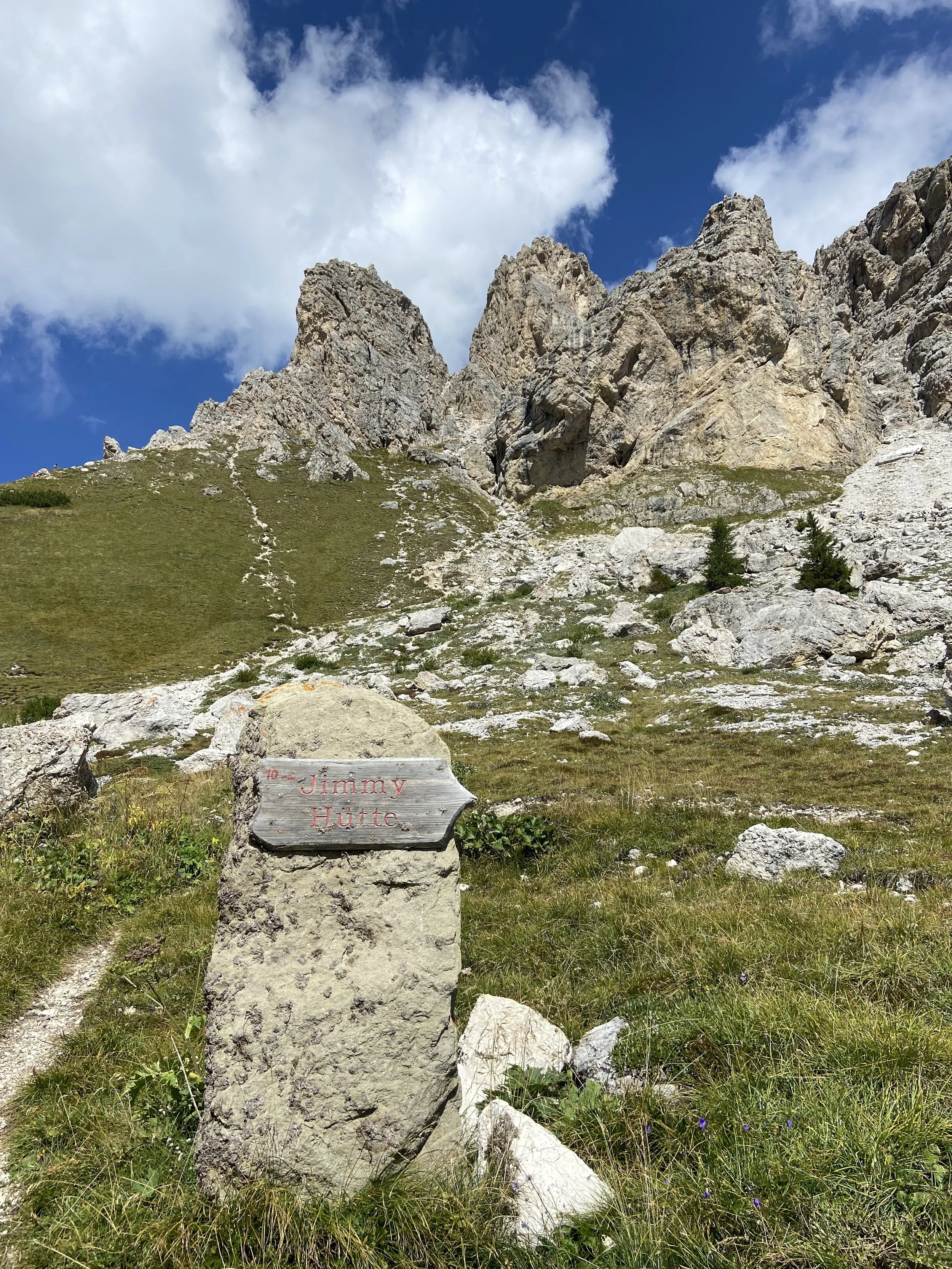 A mountain landscape with rocky peaks, green grassy slope, a trail, and a sign that reads 'Jimmy Hütte' on a stone
