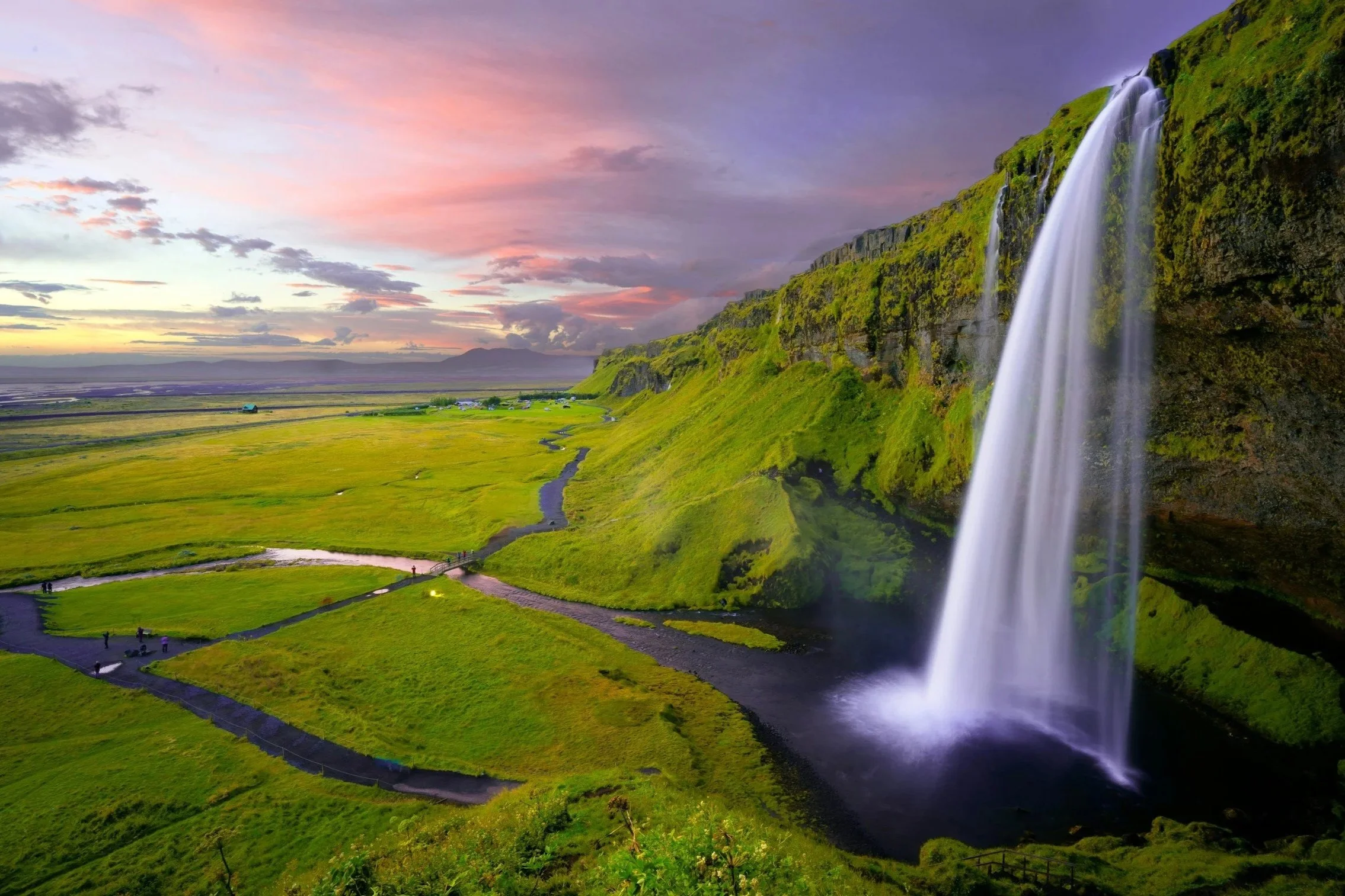 Scenic view of a waterfall cascading down a green cliff into a pond, with a lush grassy landscape and a colorful sunset sky in the background.
