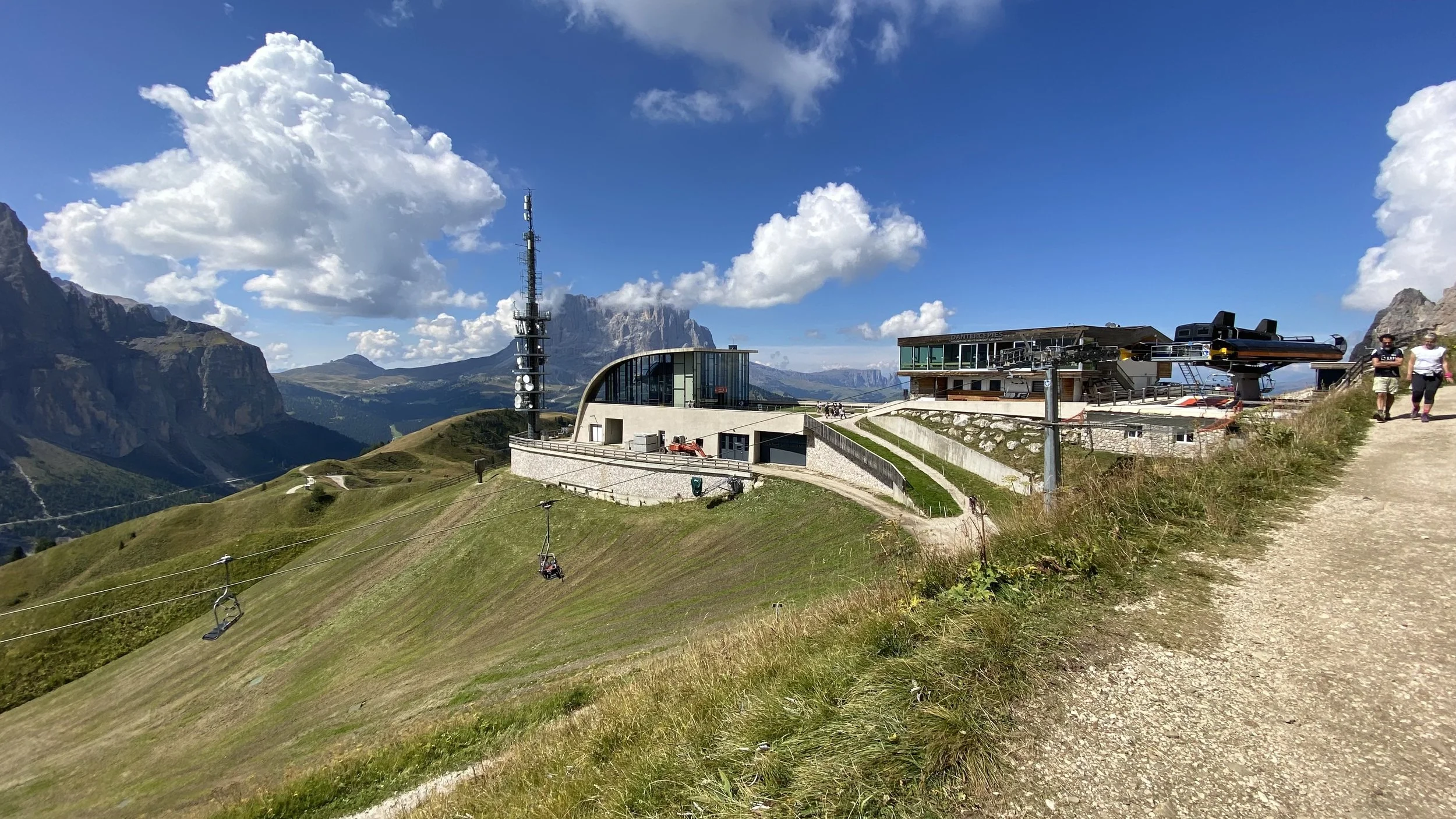 A mountain resort with modern buildings, a ski lift, and a ski slope in a lush valley under a partly cloudy sky, with mountains in the background and two people walking on a dirt path.