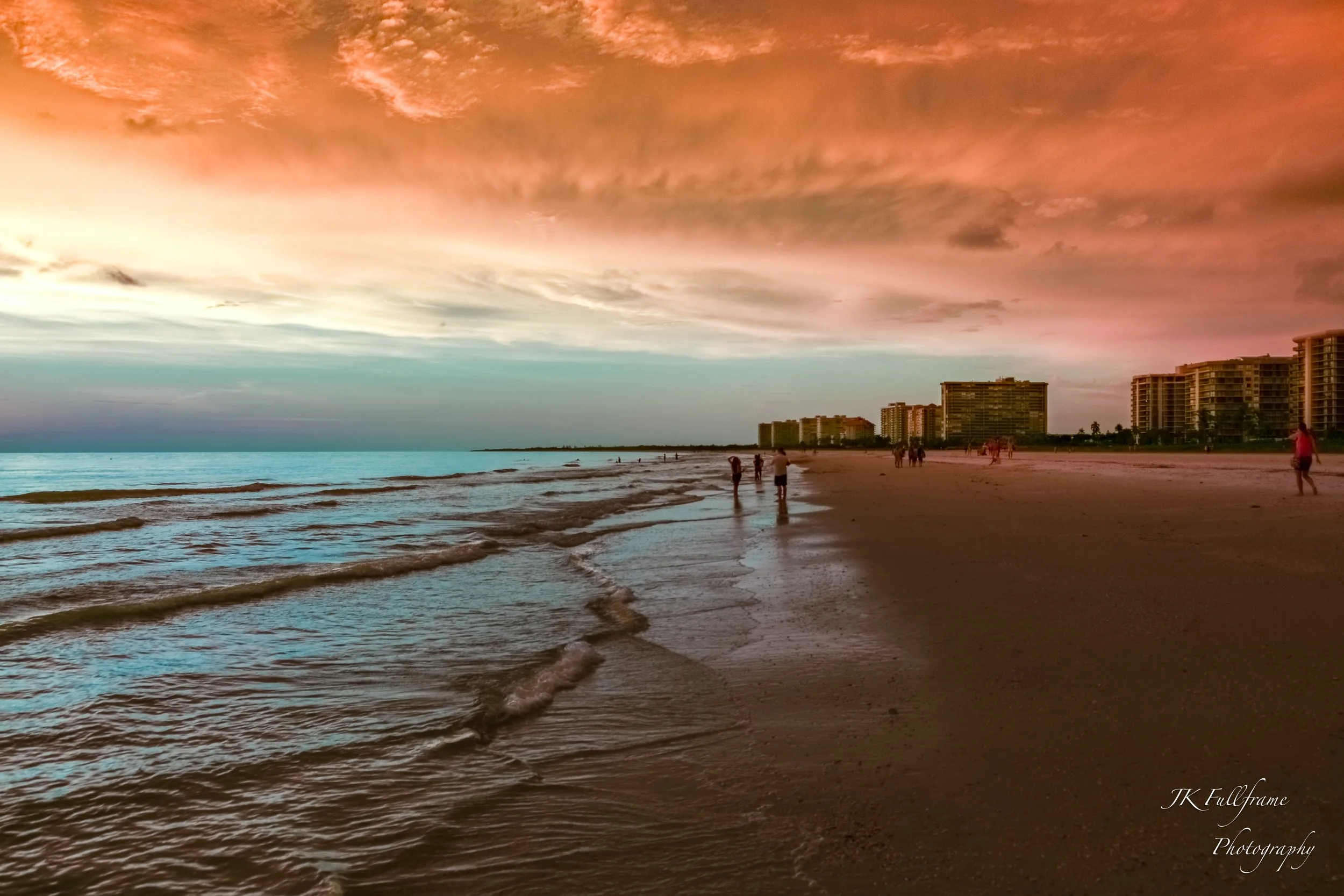 A sunset over a beach with a colorful sky, gentle waves, and people walking along the shoreline. There are high-rise buildings in the background.