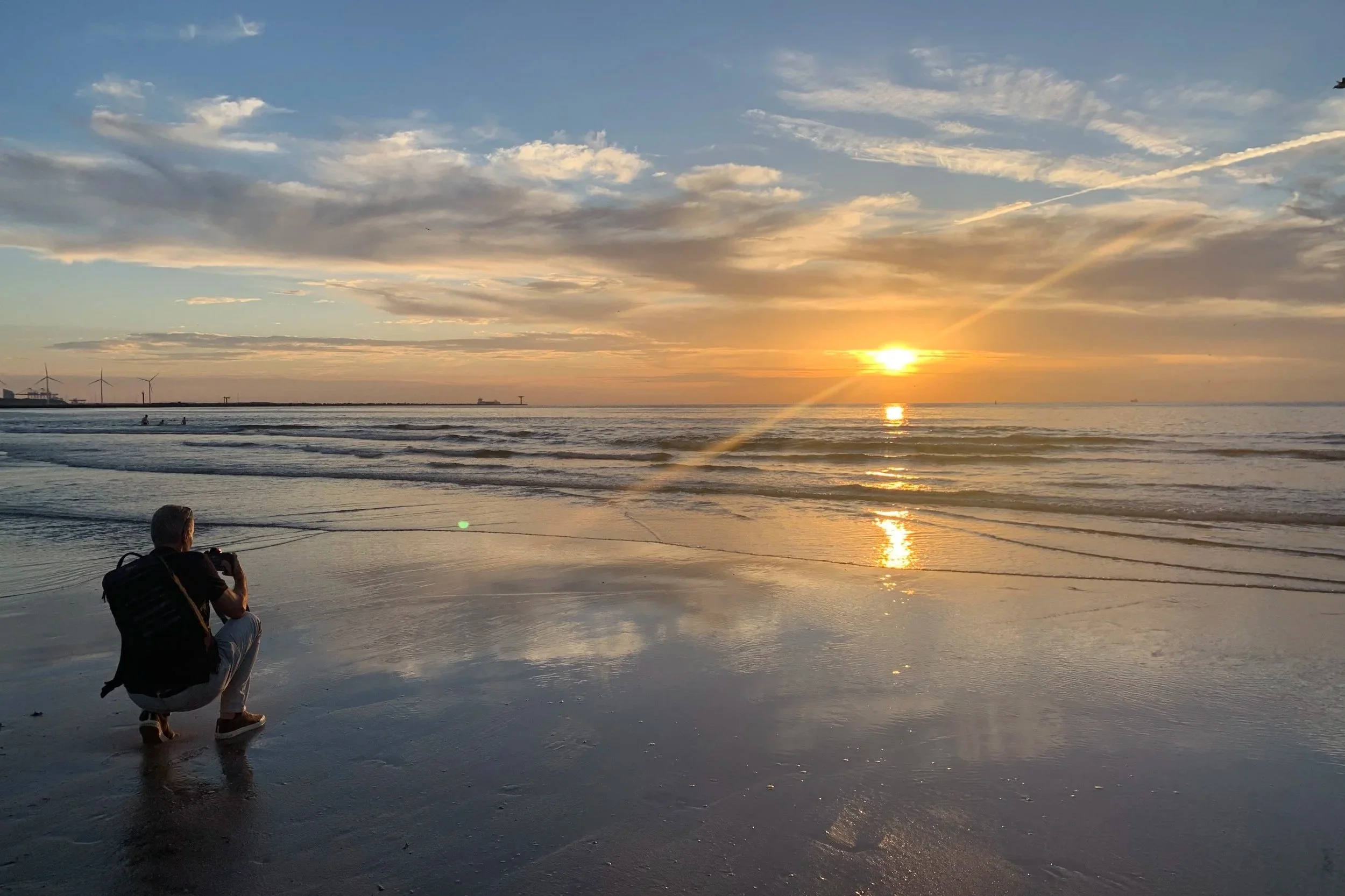 Person crouching on wet sandy beach, taking photos of the sunset over the ocean with clouds in the sky.