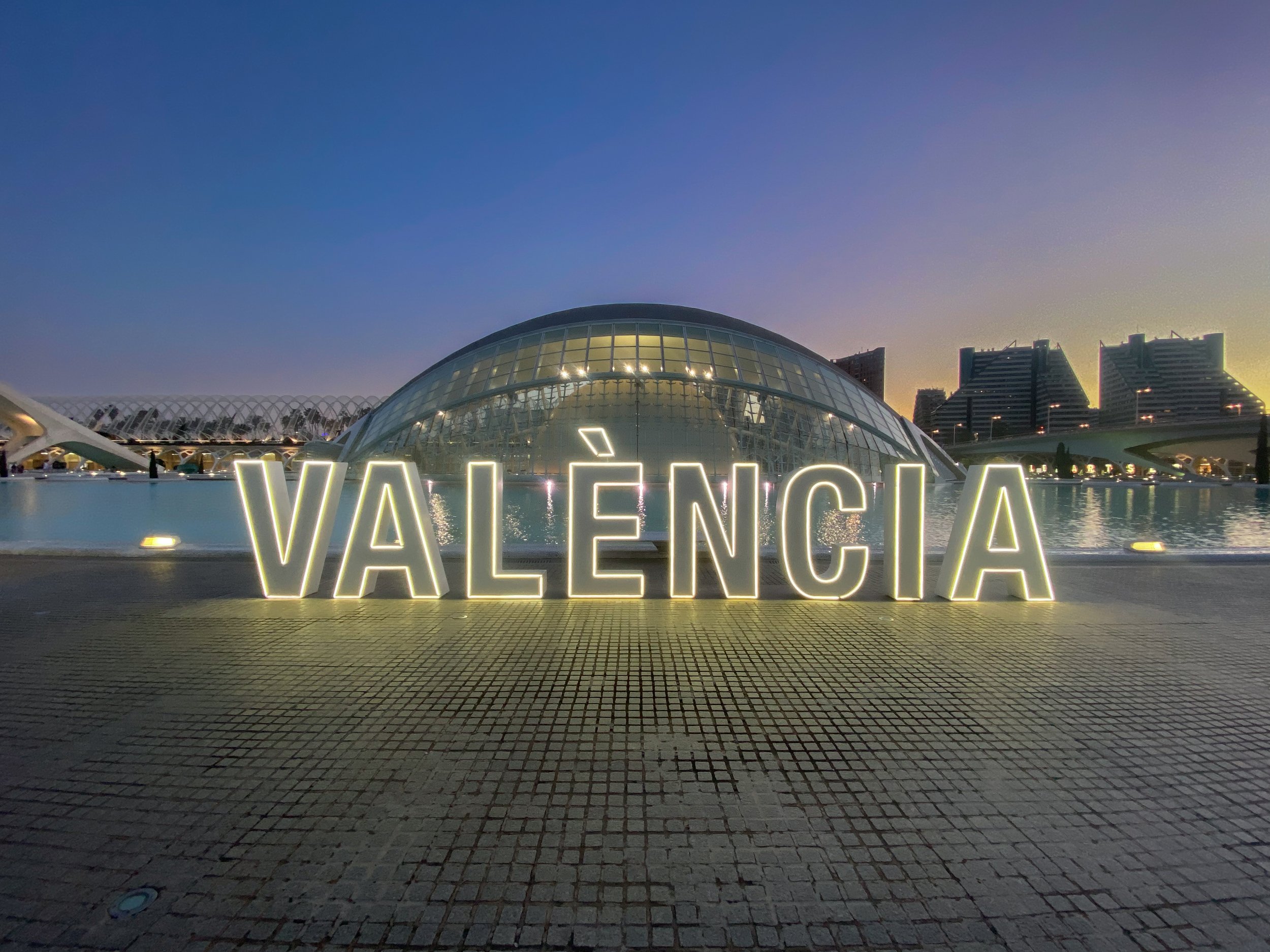 Nighttime view of the City of Arts and Sciences in Valencia, Spain, with illuminated signage spelling 'VALENCIA' in the foreground, modern architectural structures in the background, and a reflective water pool.