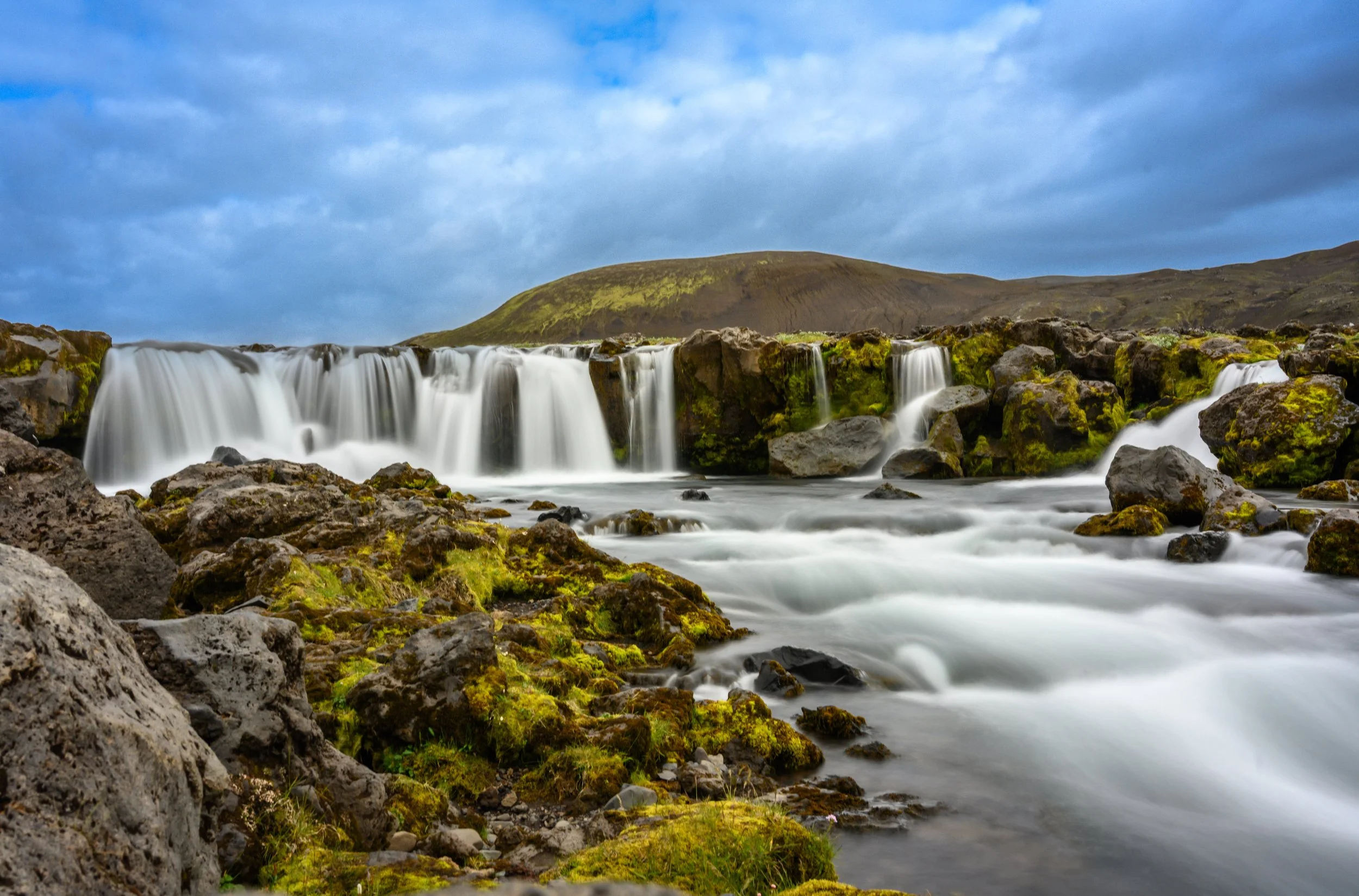 Rocky landscape in Iceland on route 210 with a serene waterfall cascading over mossy rocks into a clear stream. Green grass in the foreground; overcast sky above. Peaceful ambiance.