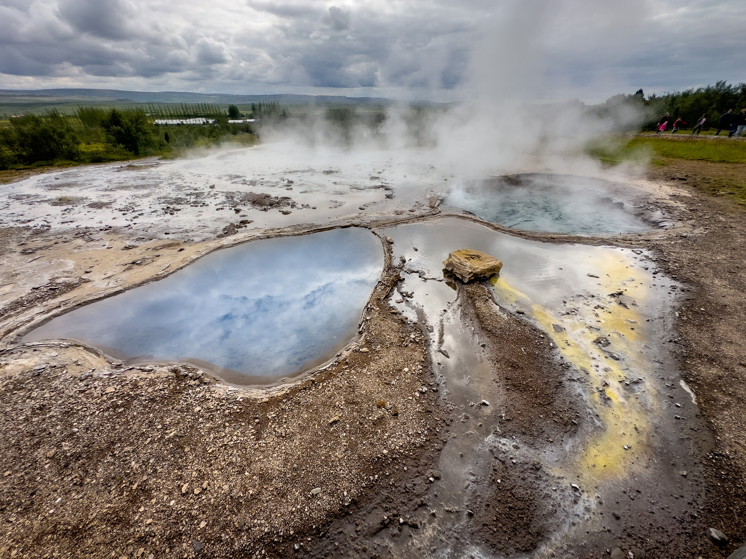 A dome of turquoise water swells and then erupts from the Strokkur geyser in Iceland, sending a tall plume of white steam and water skyward, with onlookers gathered around the geothermal pool in Iceland's Haukadalur valley