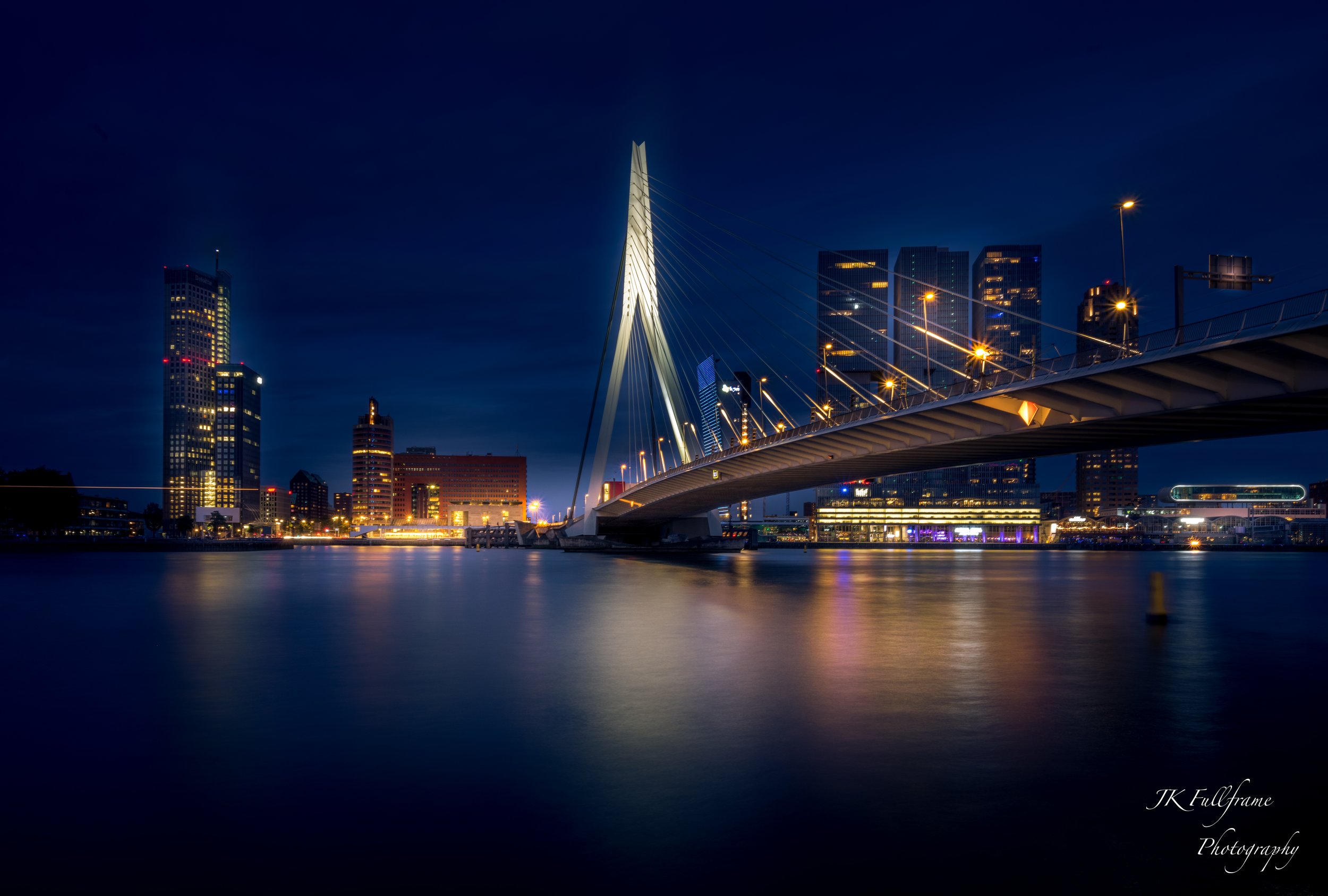 Night view of a modern city skyline featuring a distinctive cable-stayed bridge over water, with illuminated skyscrapers and buildings in the background.