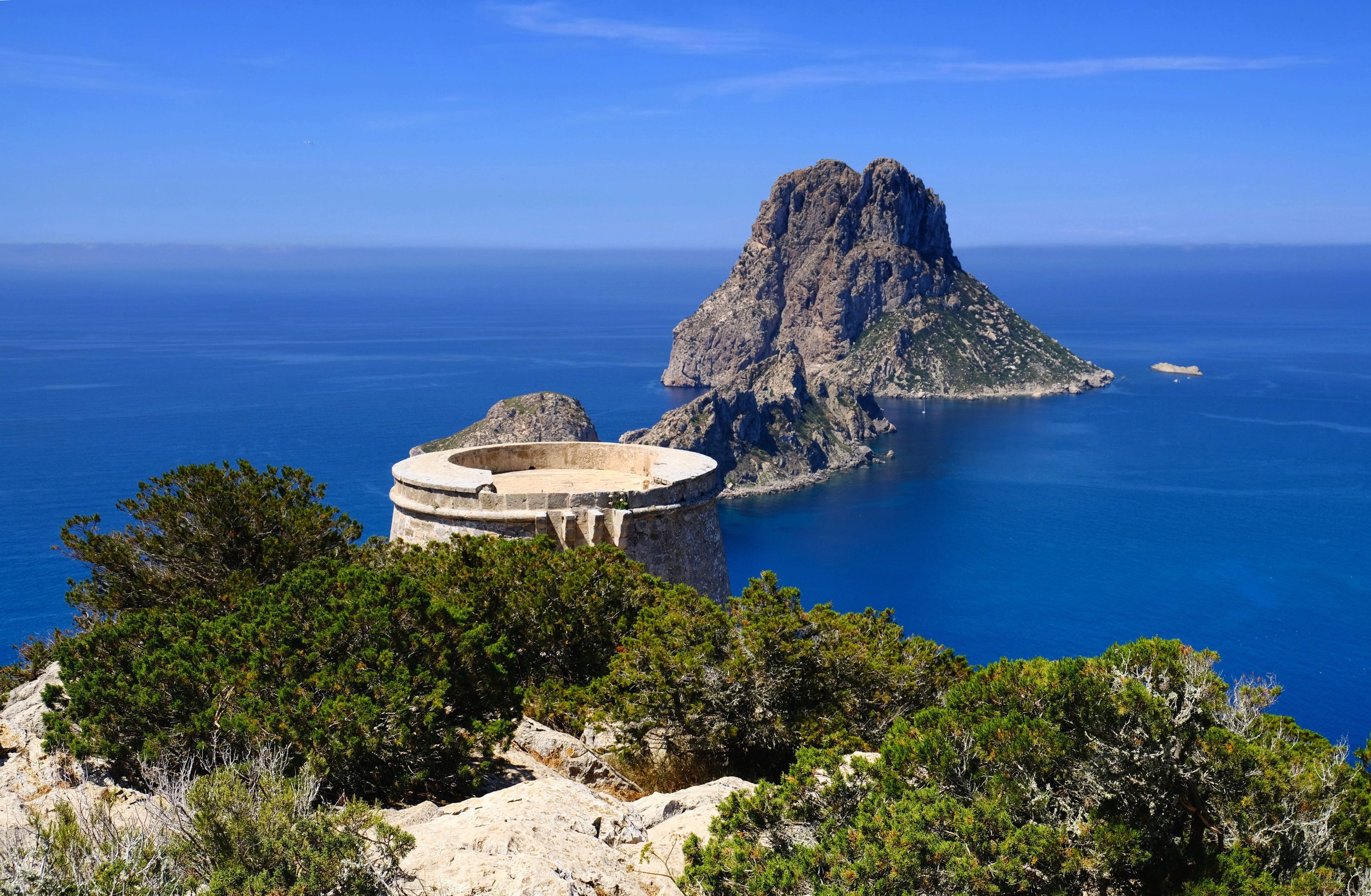 A coastal landscape with a rocky island in the sea, a blue sky, and a stone observation tower surrounded by green shrubs in the foreground.
