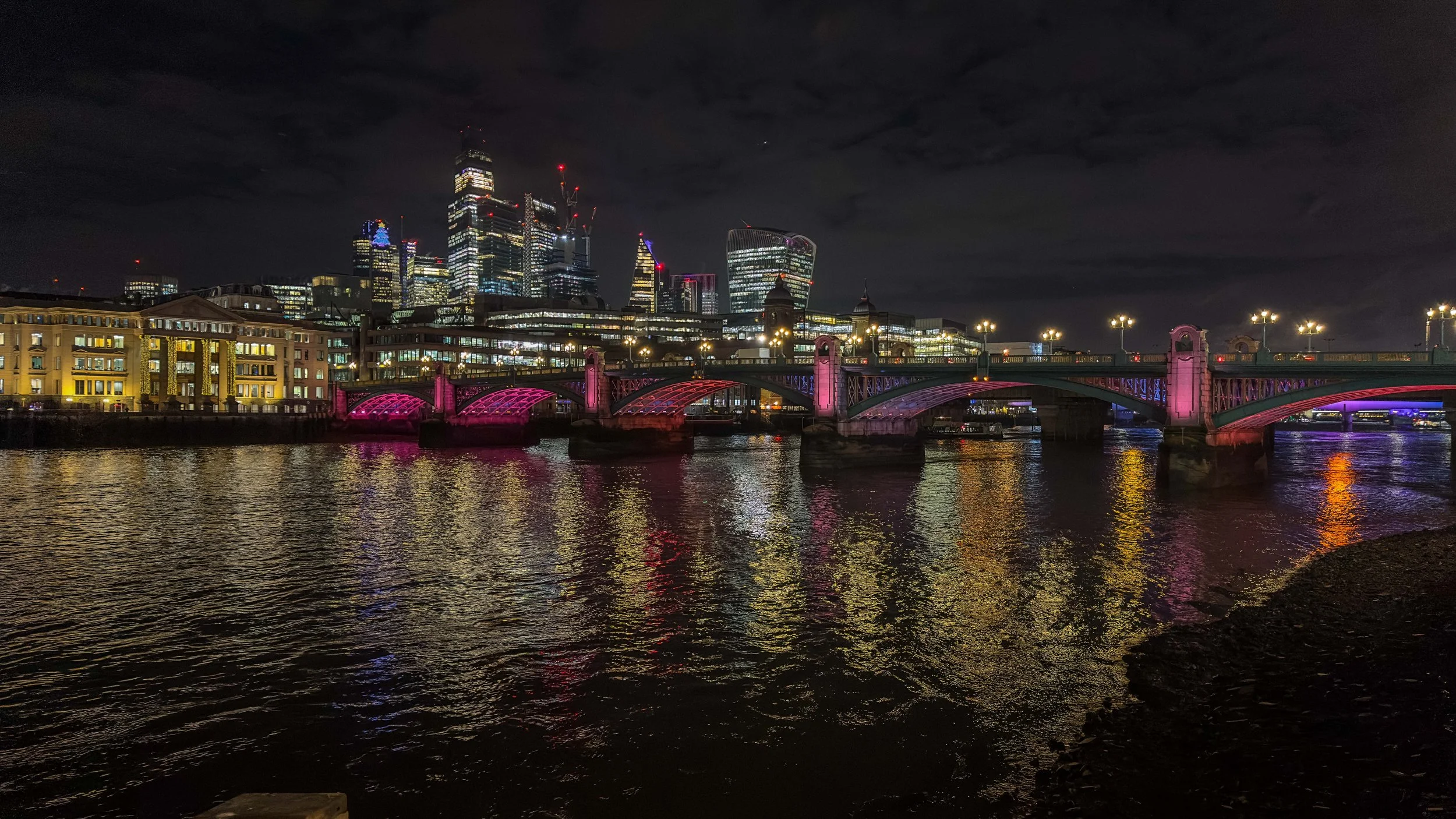 Night view of a city skyline with tall buildings and a bridge illuminated with pink and purple lights over a river.