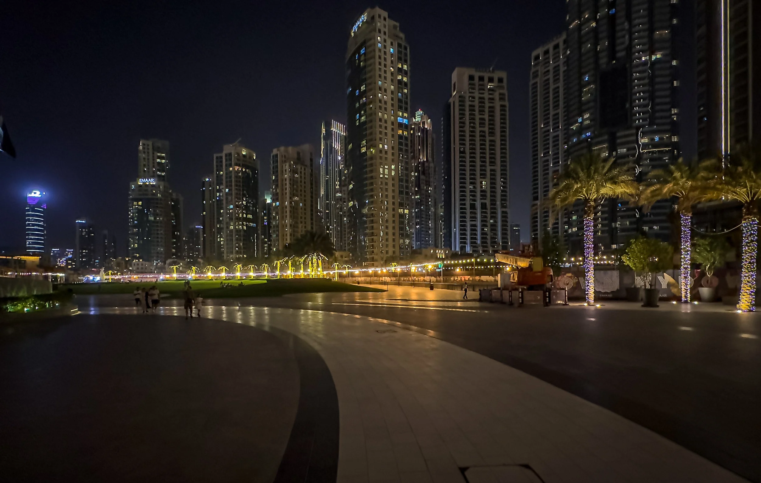 Night view of a city skyline with tall illuminated buildings and palm trees decorated with lights, along a promenade with a few people walking.