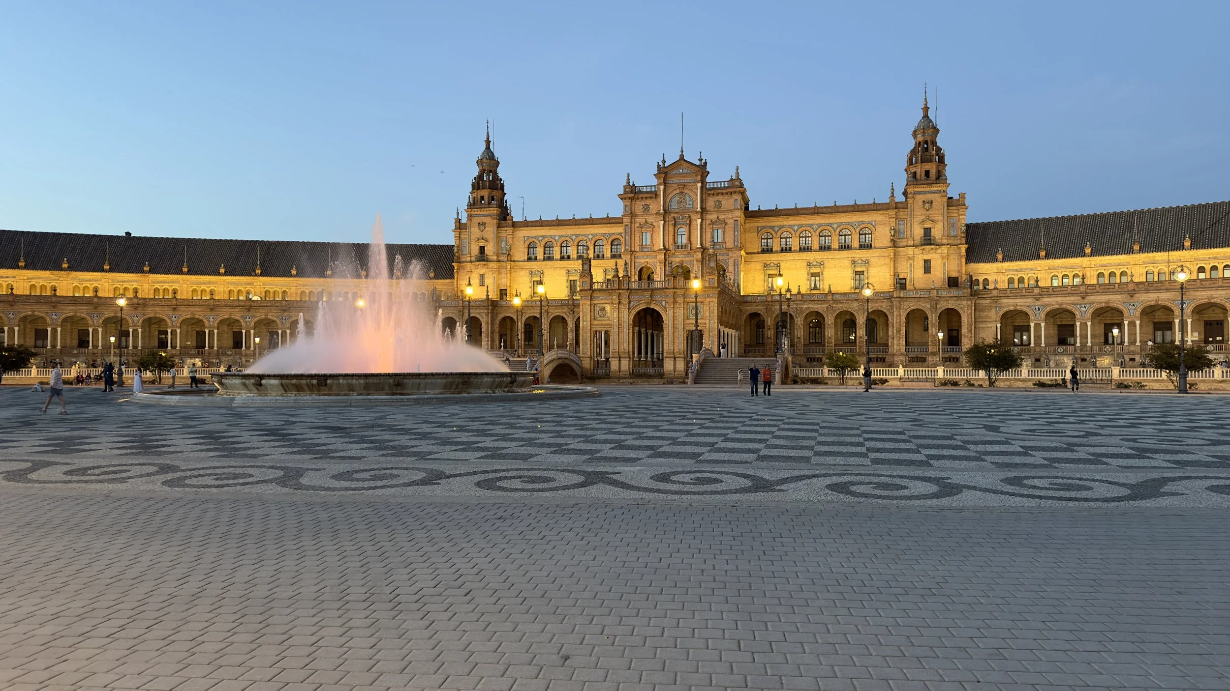 The Plaza de España in Seville, Spain, showing a large golden yellow and ornate building with towers, a central fountain with pink water spray, and a patterned stone plaza at dusk.