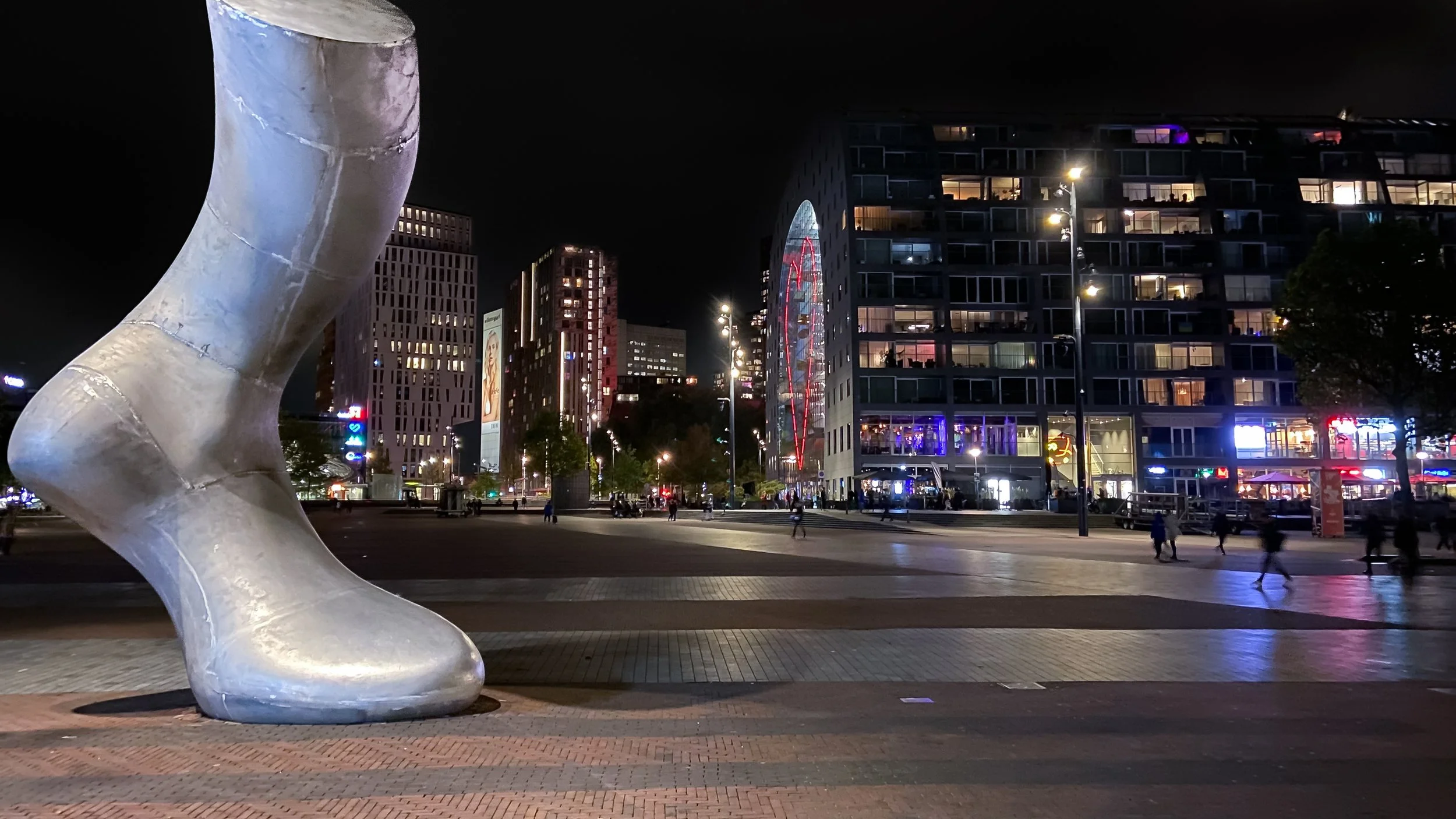 Night view of a city square with a large, silver high-heeled shoe sculpture in the foreground and modern buildings with colorful lights in the background.