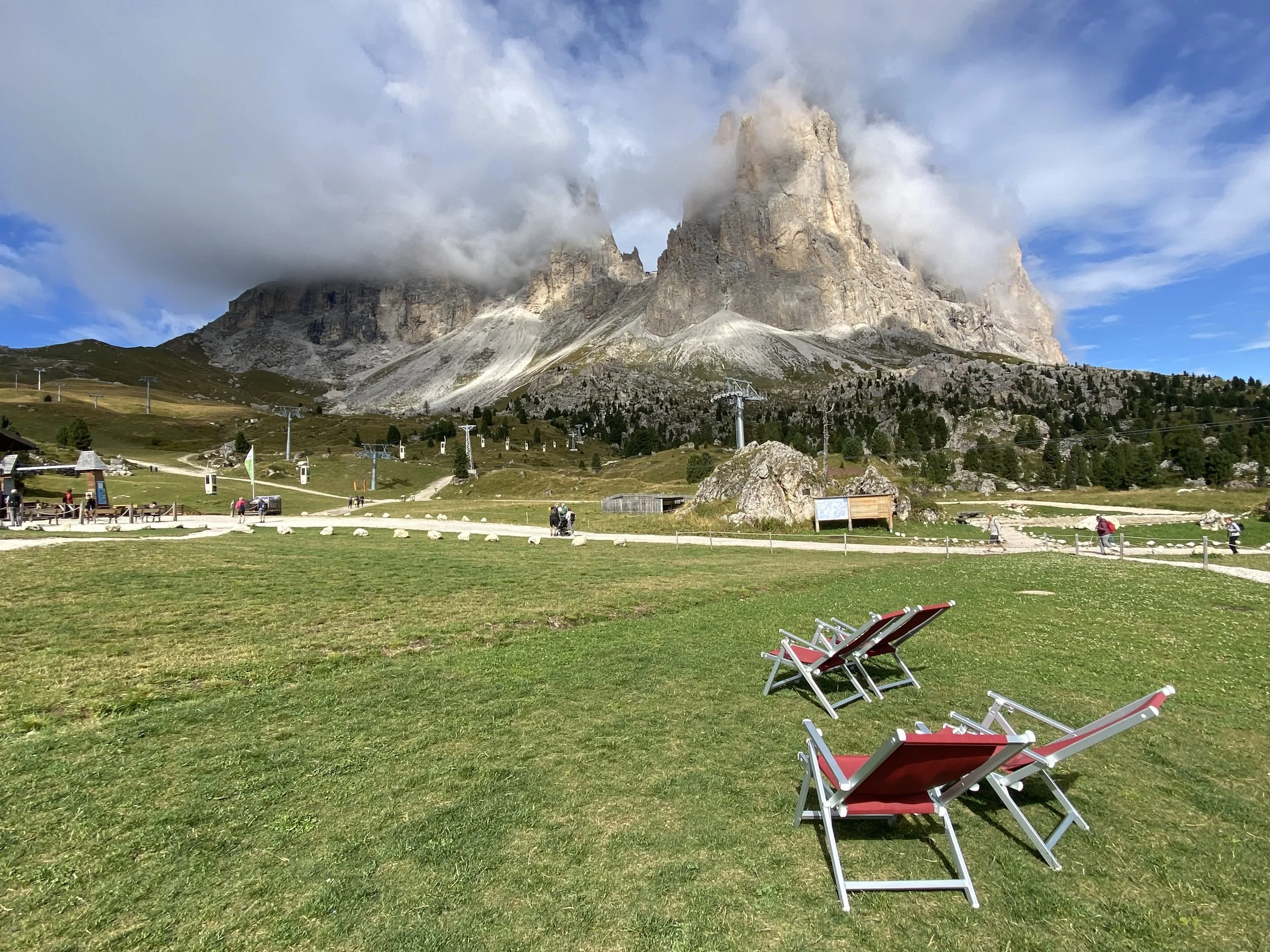Two red lounge chairs on a green grassy field with mountains partly covered by clouds in the background, and a clear sky with a few clouds.