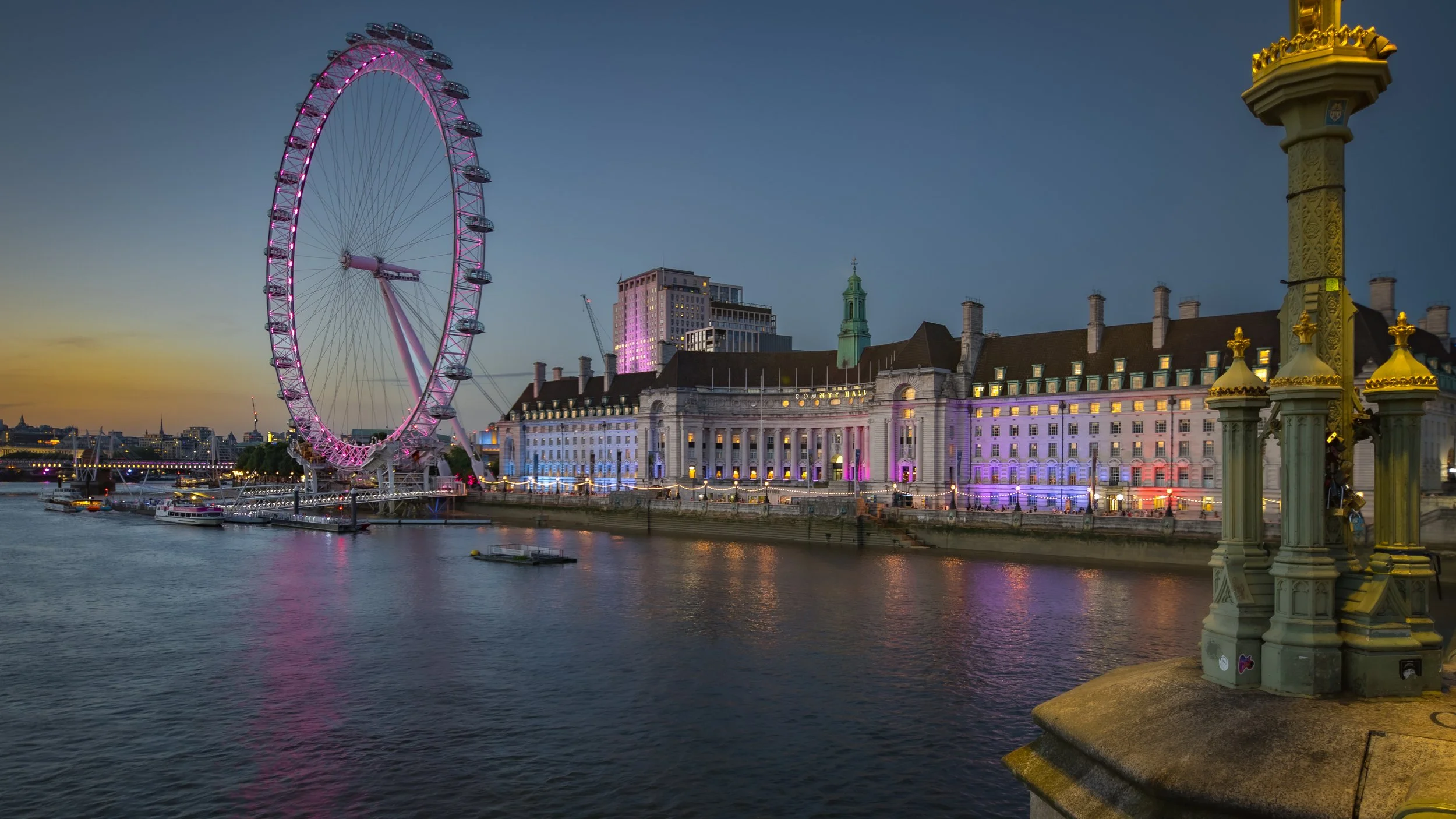 A cityscape view of the London Eye illuminated with purple and pink lights on the River Thames during dusk, with historic and modern buildings in the background.