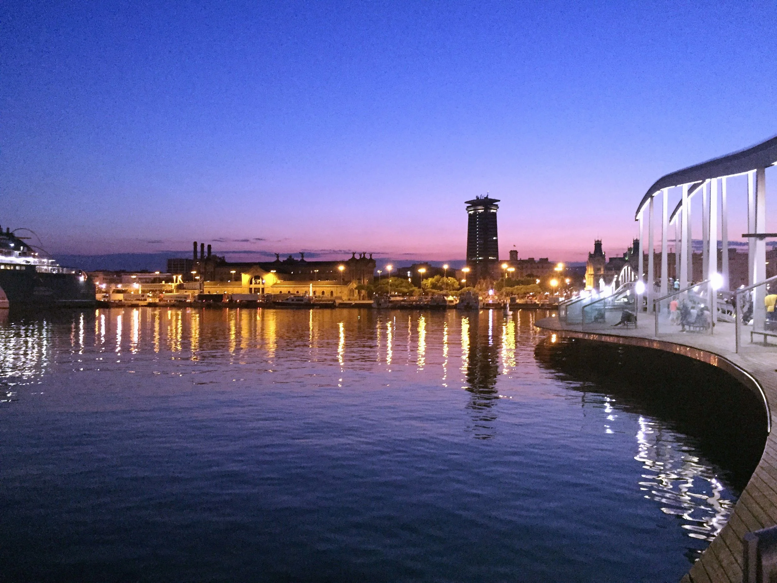 A waterfront cityscape at dusk with calm water reflecting lights from buildings, a boat on the left, and a modern curved structure with seating on the right.