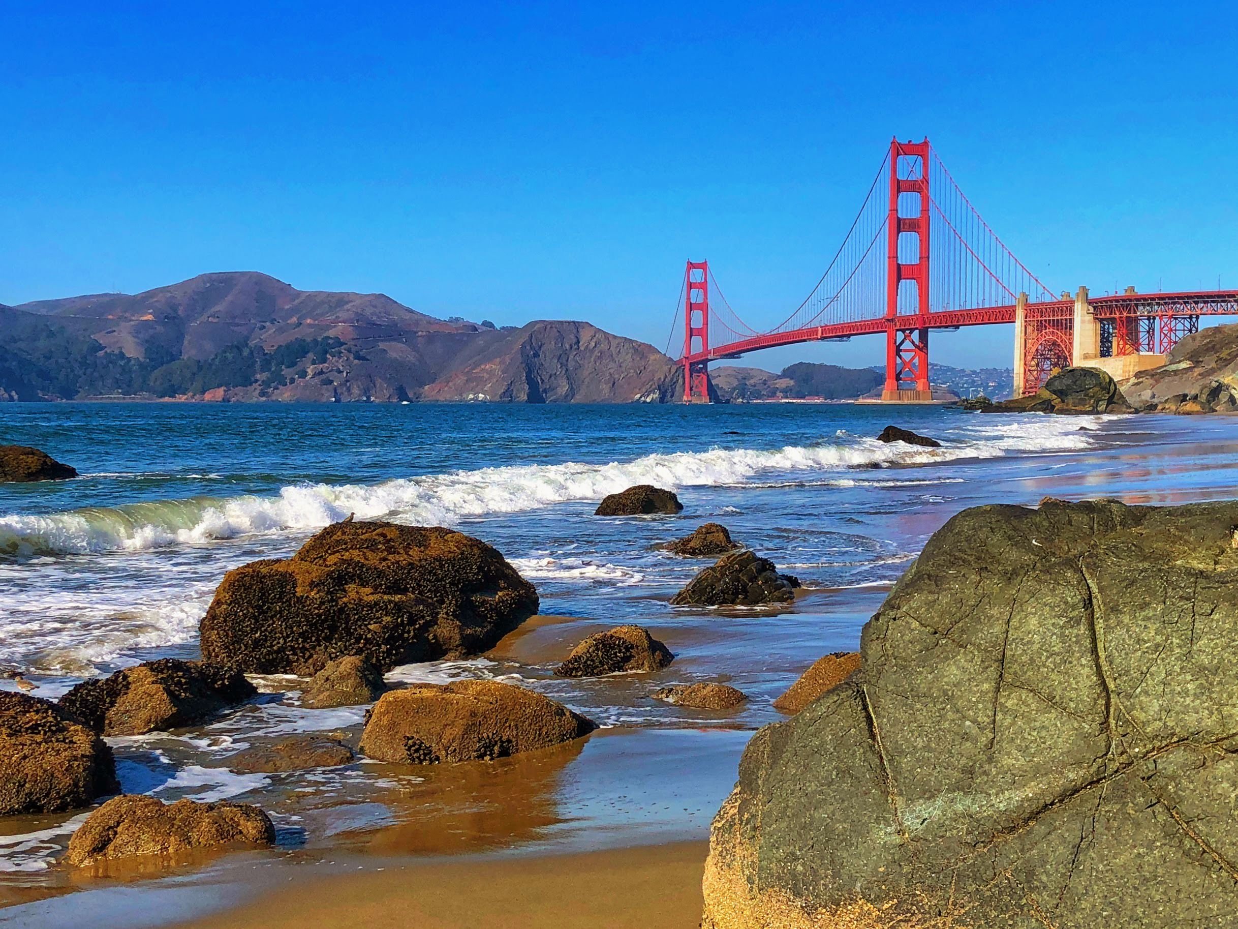 View of the Golden Gate Bridge over the water with rocks and sandy beach in the foreground and hills in the background under a clear blue sky.
