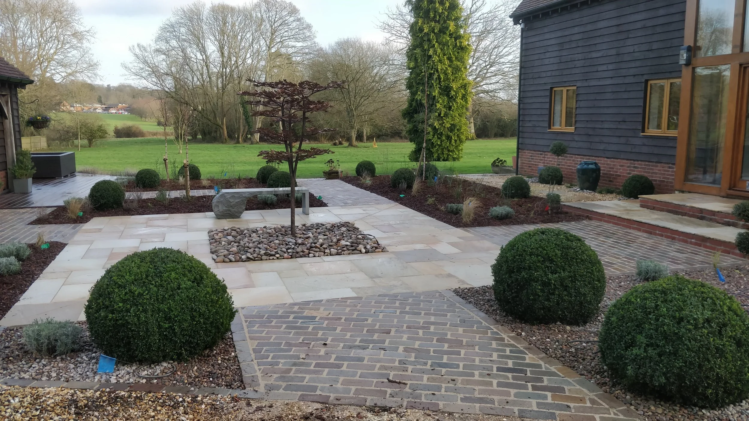 A landscaped backyard garden with a paved patio, neatly trimmed bushes, small trees, and a green lawn in the background.