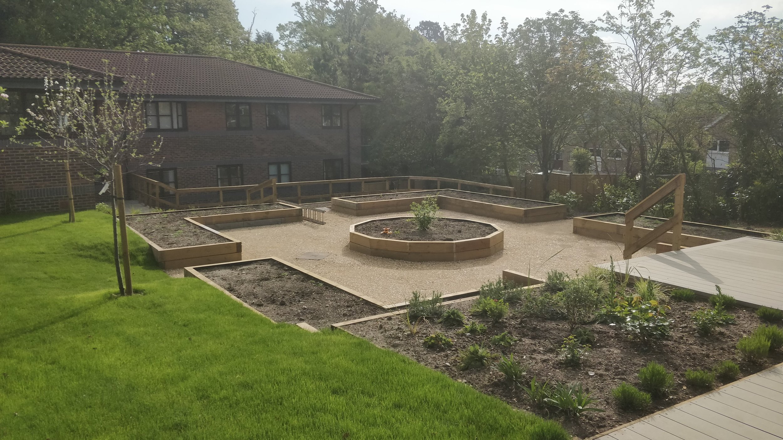 Backyard with newly built raised garden beds, small trees, and a green lawn. A deck with a staircase is visible on the right, and there is a brick building in the background surrounded by trees.