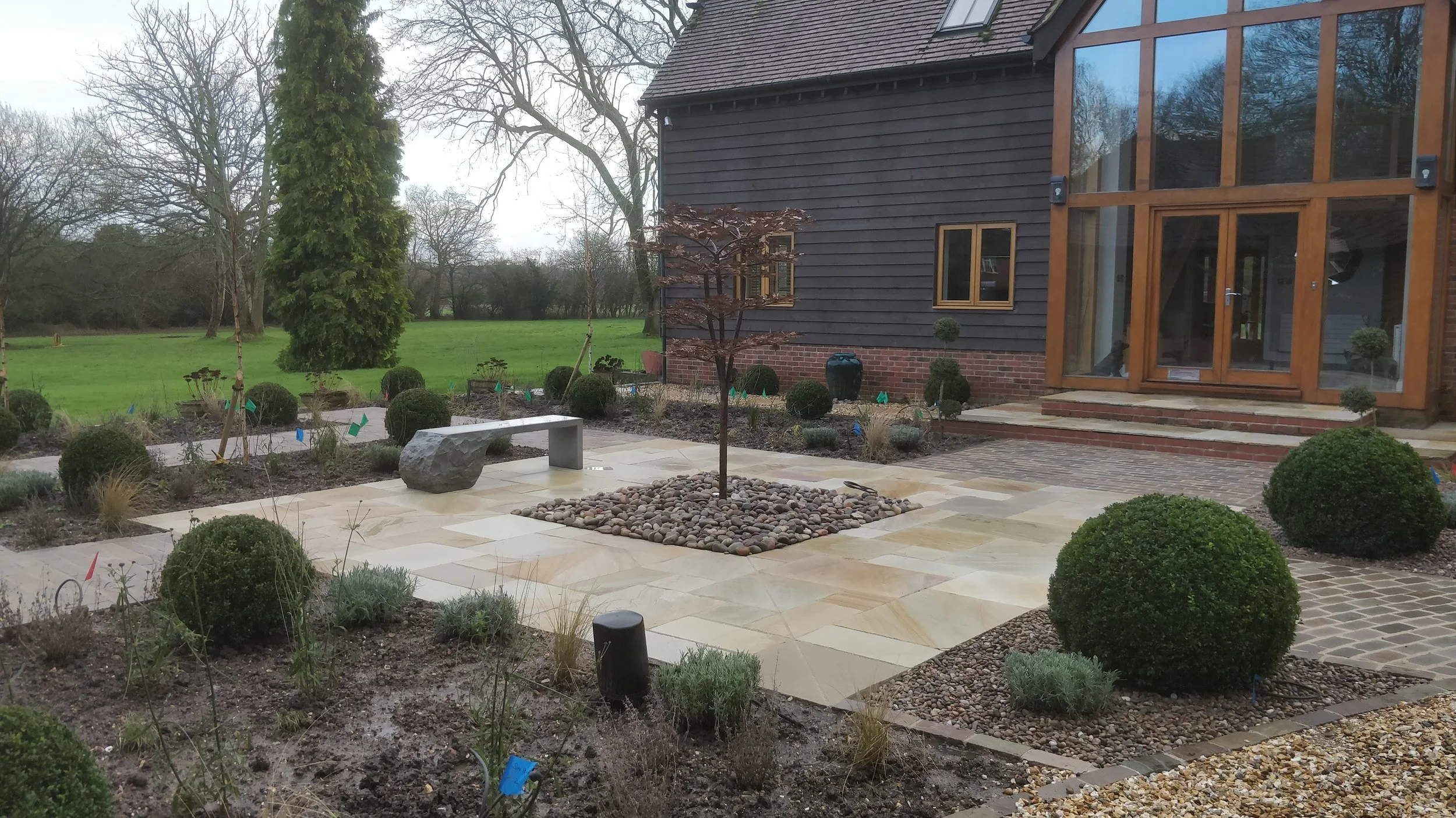 A newly landscaped backyard patio with a central young tree surrounded by small rounded stones. The patio has large light-colored stone tiles and includes a stone bench. Nearby are round, neatly trimmed bushes and small plants in garden beds, with a 