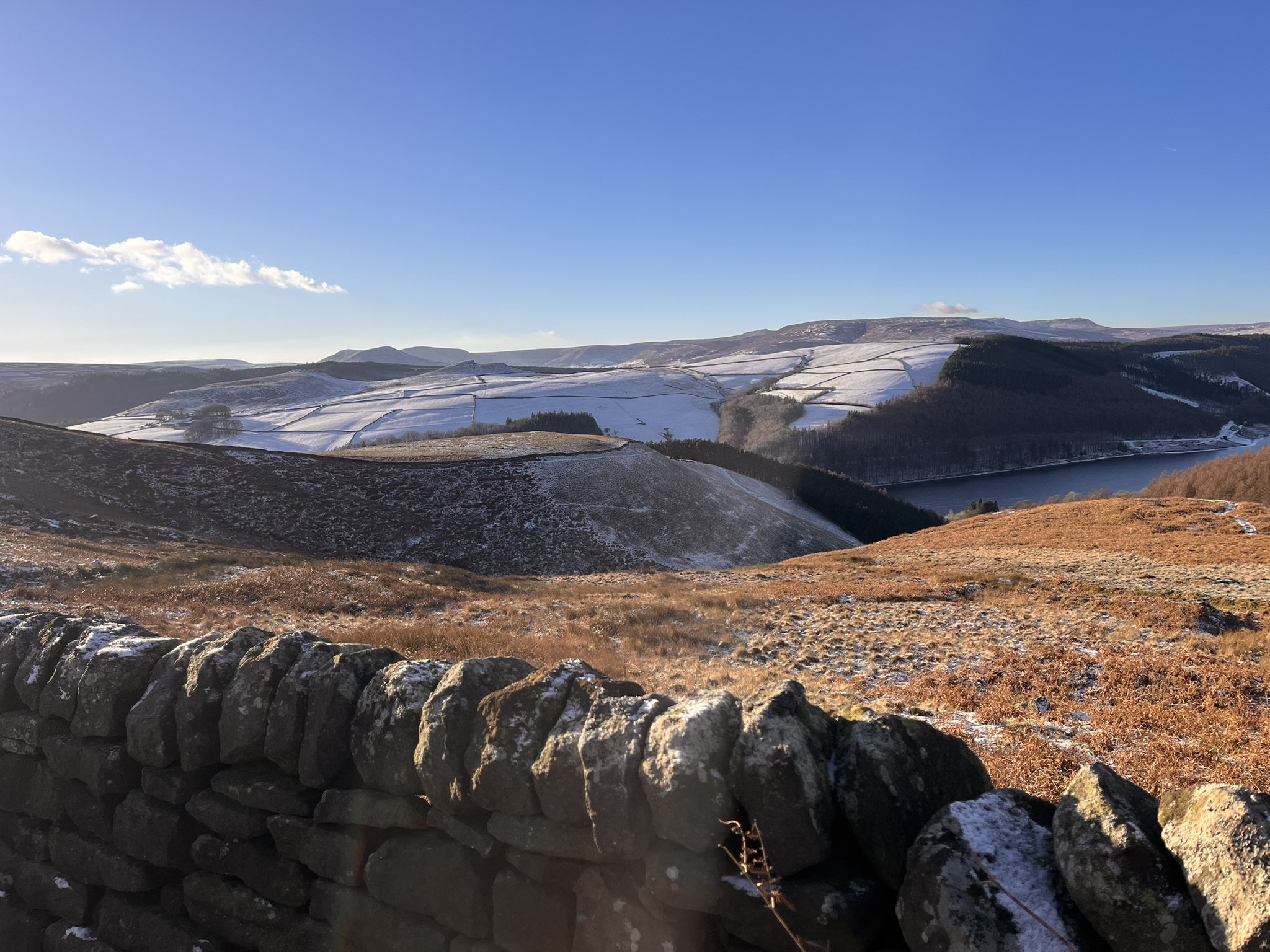 Snow-capped hills and valleys of a Peak District view under a clear blue sky, with a stone wall in the foreground.