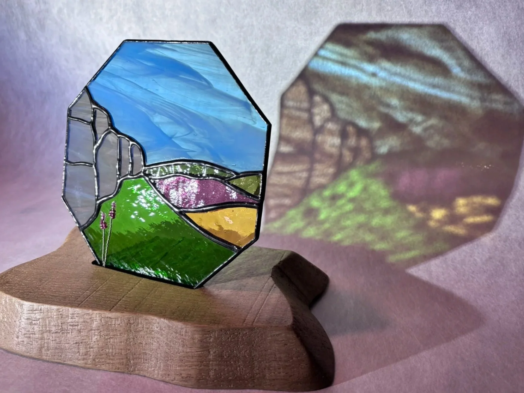 A stained glass artwork of a landscape scene featuring a blue sky, mountains, fields, and trees, placed on a wooden stand, casting a colorful shadow on the background wall.