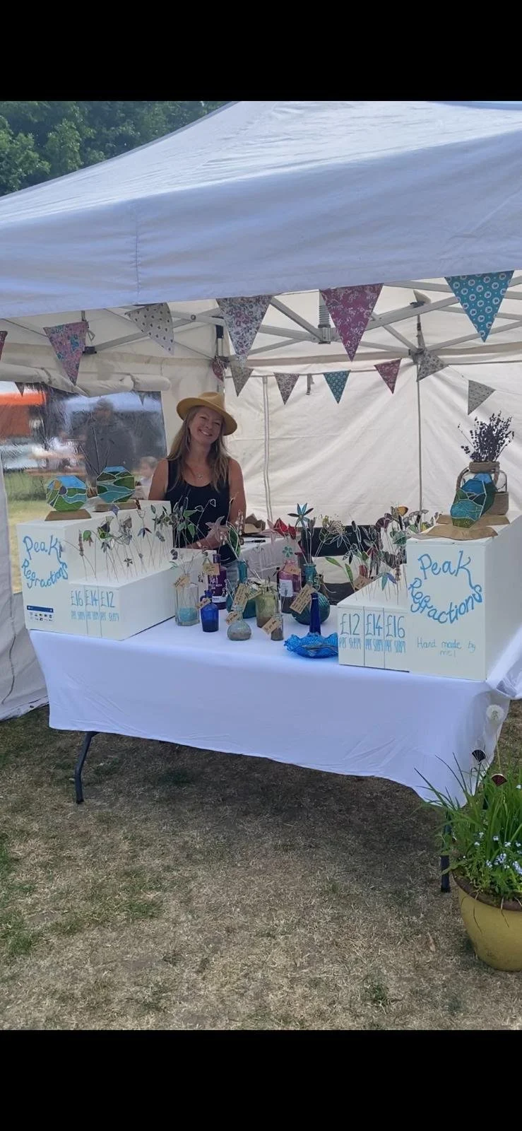 Peak Refractions market stall  featuring my white gazebo decorated with colorful bunting and a table of stained glass flowers in gin bottles at a summer Sheffield outdoor market.