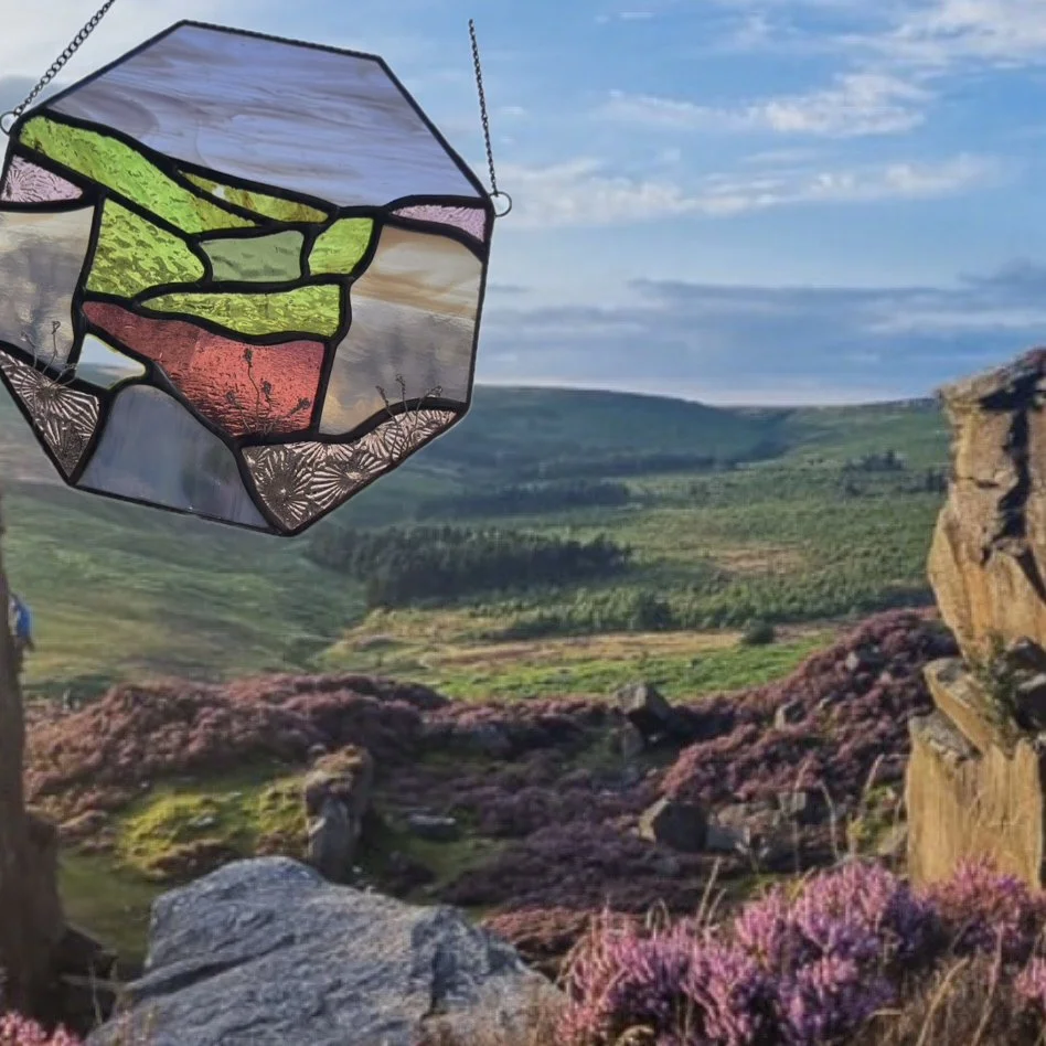 A stained glass ornament depicting a landscape with rolling hills, grass, and sky, hanging outdoors against a scenic background of green hills, purple heather, rocks, and blue sky with clouds.