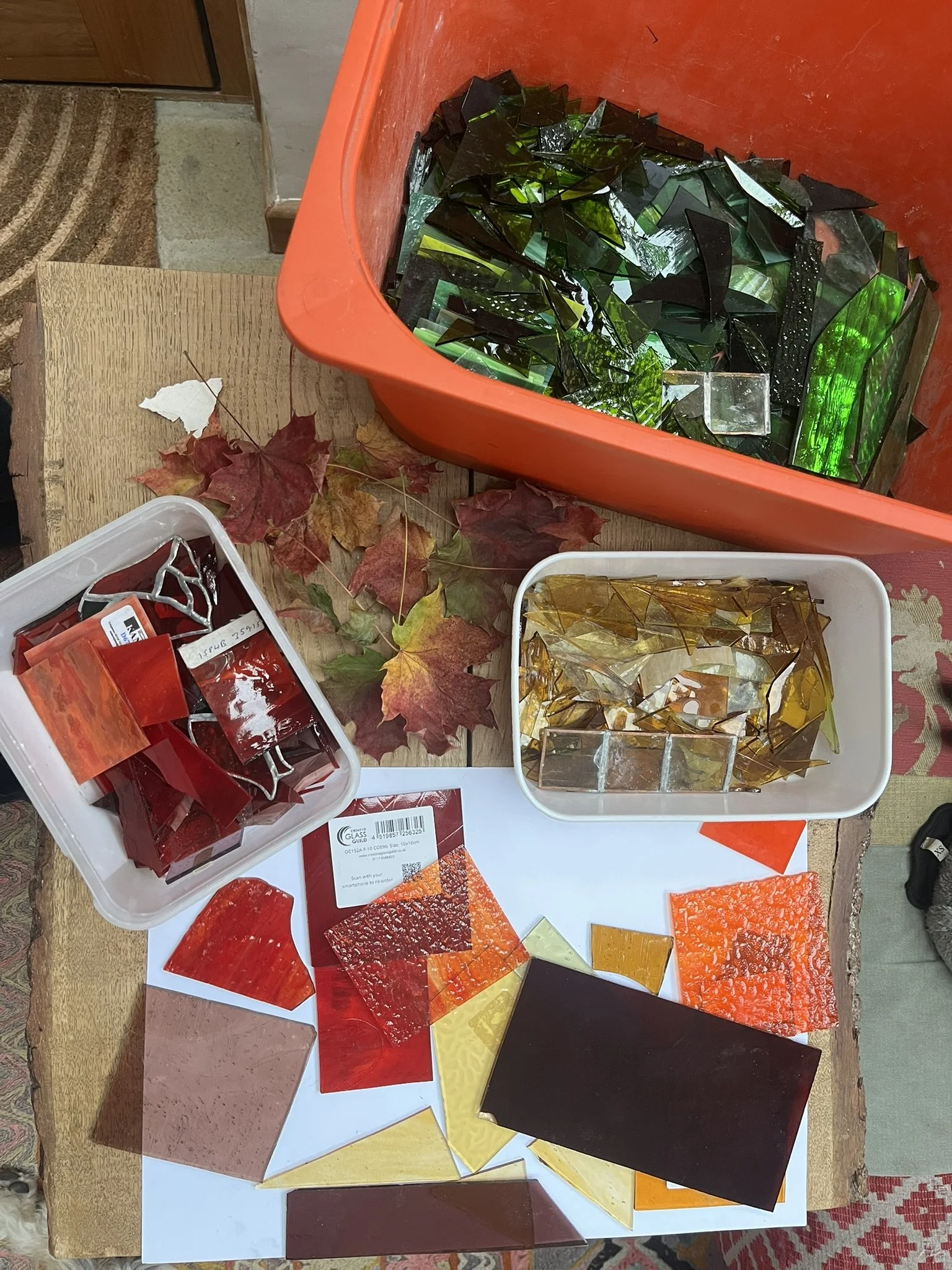 Colorful broken pieces of stained glass in various shades of red, orange, yellow, green, and black, arranged on a white paper and in containers, along with some autumn leaves on a wooden table.