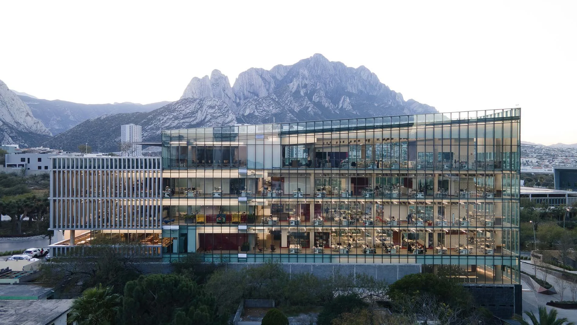 Edificio moderno de varios pisos con fachada de cristal, rodeado de naturaleza y montañas en el fondo.