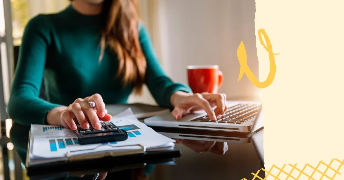Mujer trabajando con calculadora y computadora portátil en un escritorio con papeles y una taza roja.