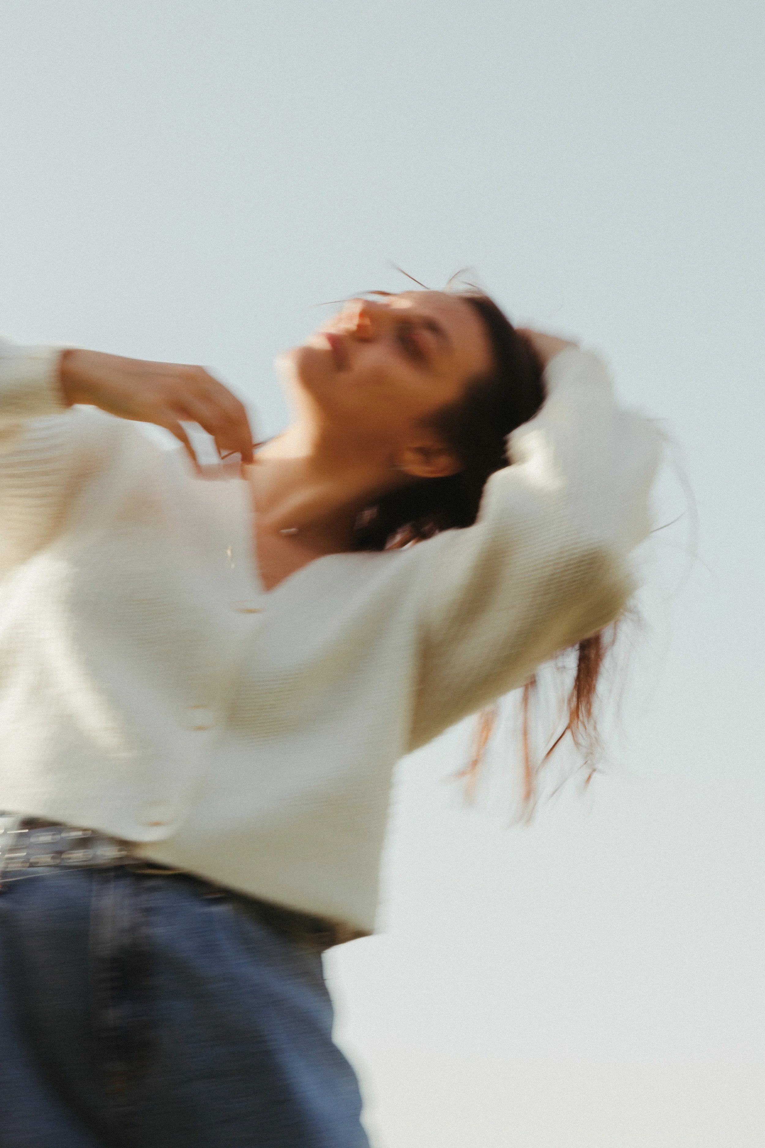 A woman wearing a cream-colored sweater and blue jeans, with her hand behind her head, looking up against a clear sky.