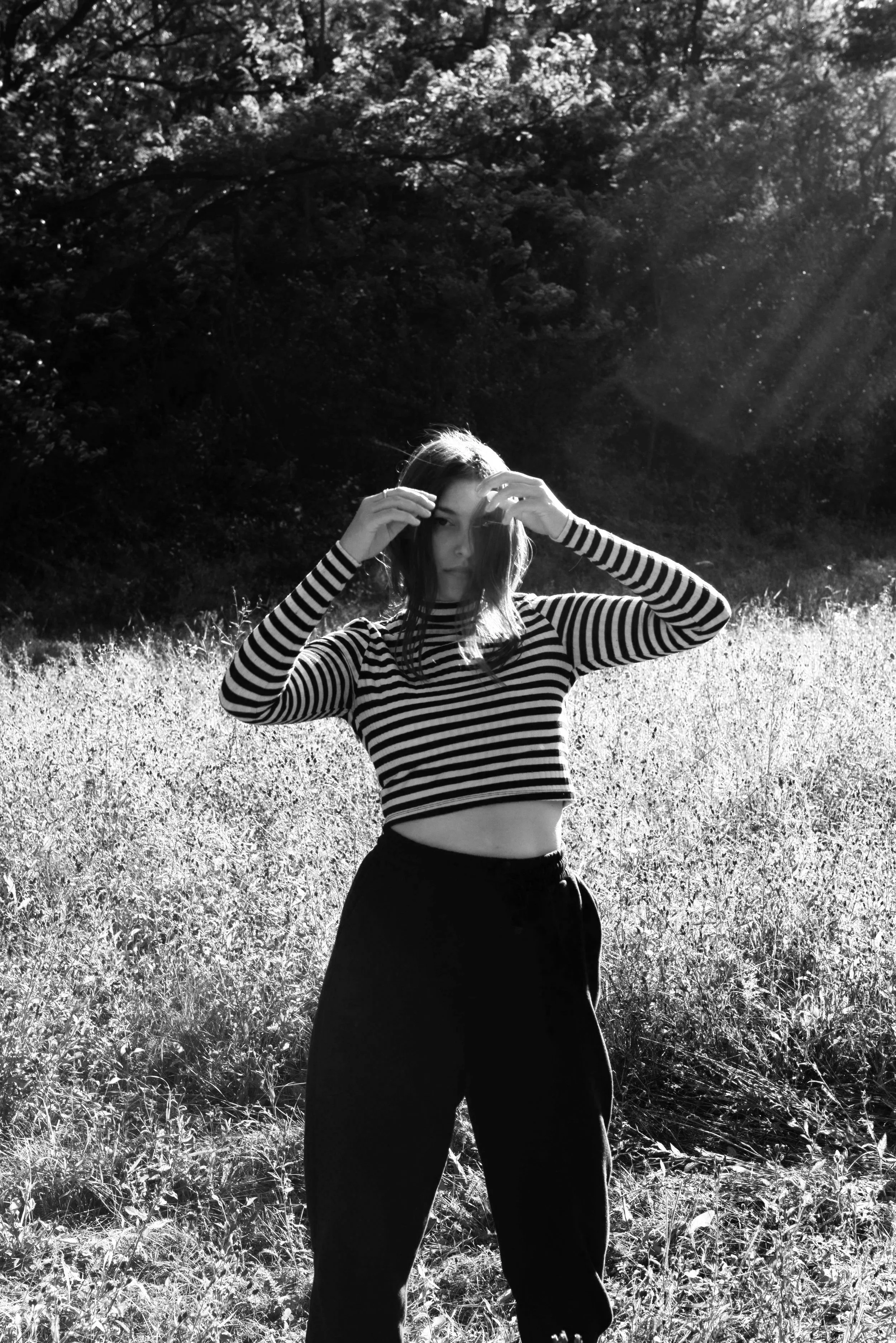 A young woman in a striped long-sleeve crop top and dark pants standing in a grassy field with trees in the background. The sunlight filters through the trees, creating a glow around her.