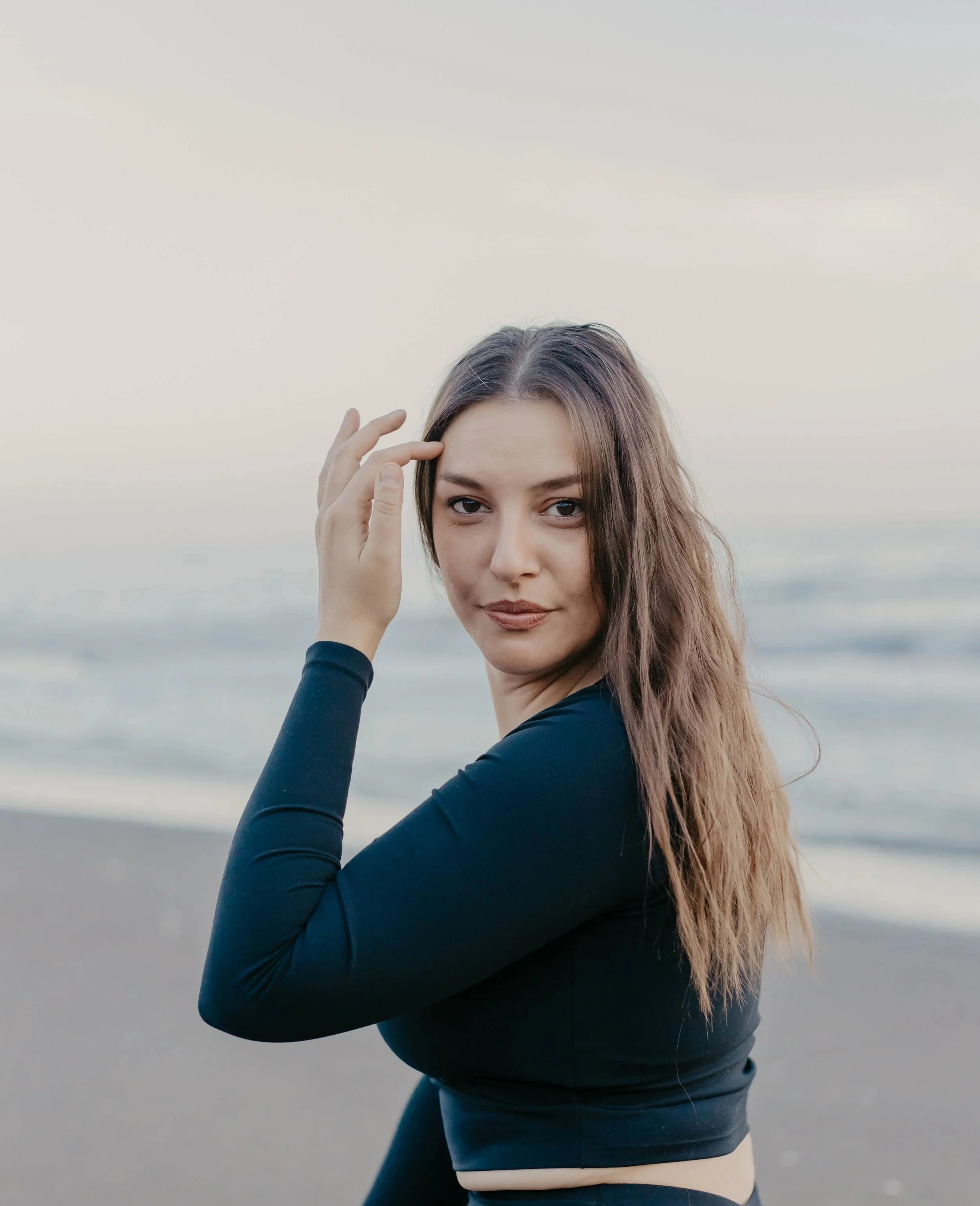 A young woman with long, wavy brown hair wearing a black long-sleeve top on a beach during sunset, looking at the camera.
