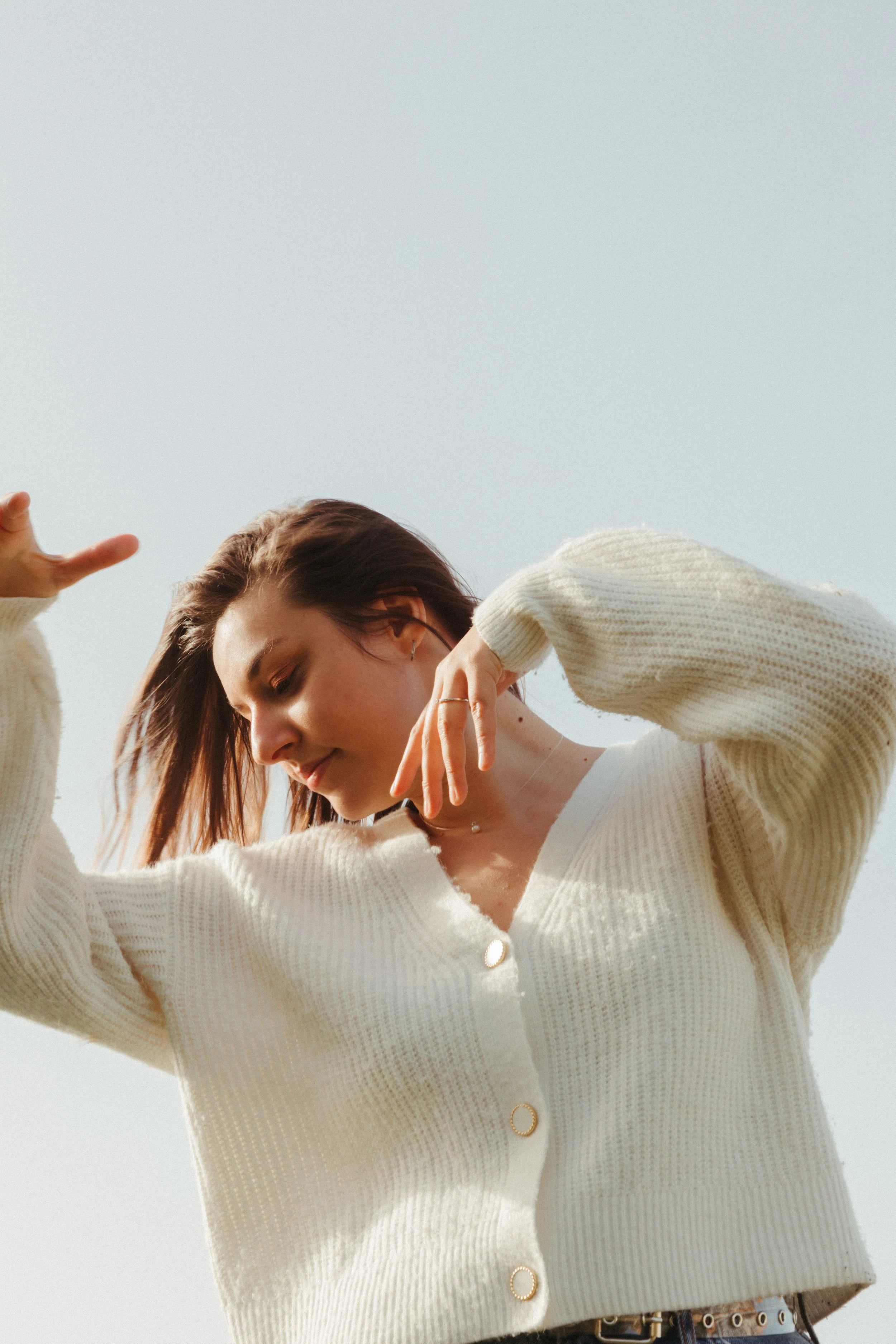A young woman with shoulder-length brown hair, wearing a cream-colored cardigan, is outdoors against a clear sky. She is looking downward with her eyes closed, smiling softly, with her left arm raised and her right hand near her face.