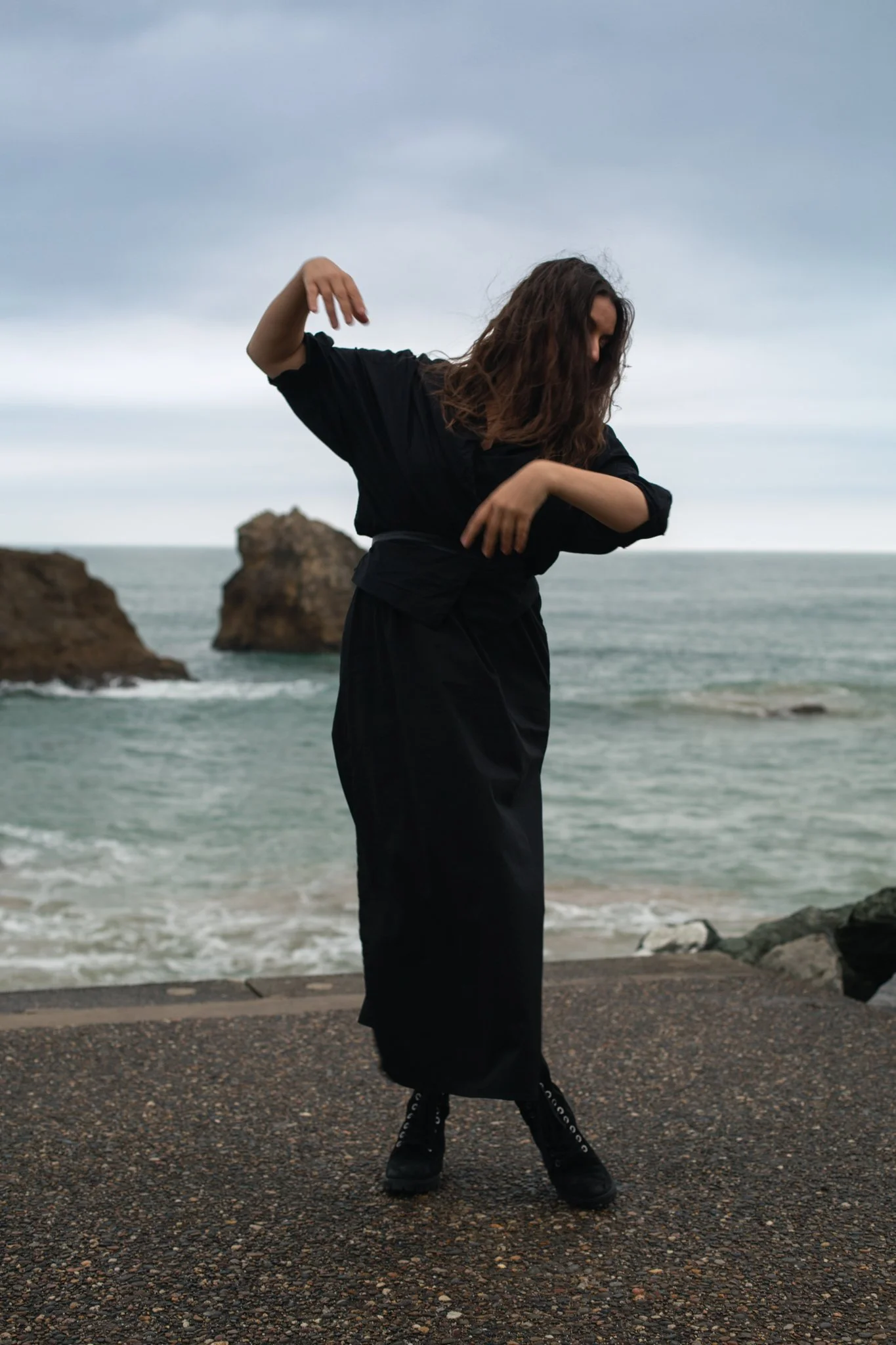 A woman dressed in black standing on a rocky surface near the ocean, with large rocks in the water and cloudy sky in the background, striking a dance pose.