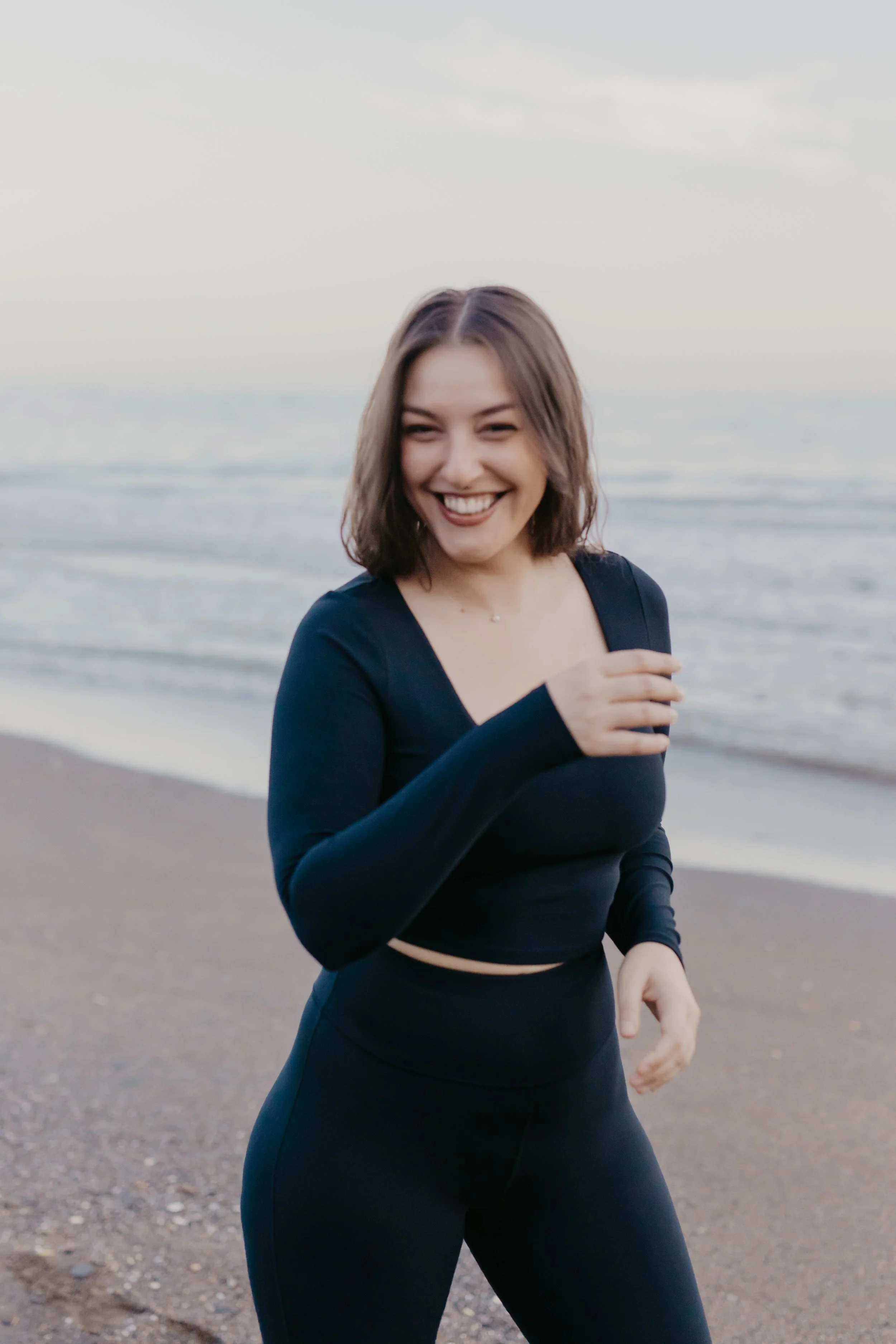 A young woman with shoulder-length hair, smiling and laughing while jogging on a beach at sunset, wearing a long-sleeve navy athletic top and black leggings.