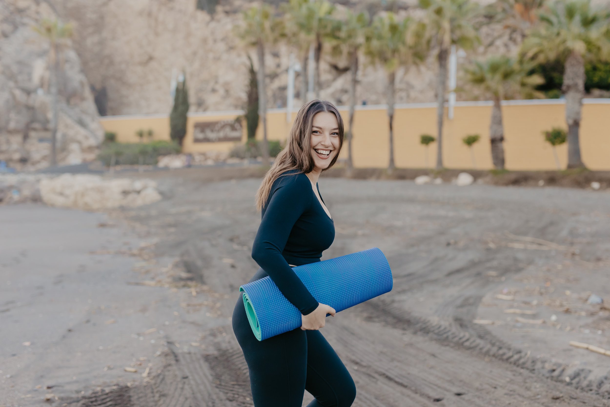 A smiling woman with long brown hair, wearing a black long sleeve shirt and leggings, is holding a rolled-up blue yoga mat while standing outdoors on a dirt path, with trees and a yellow wall in the background.