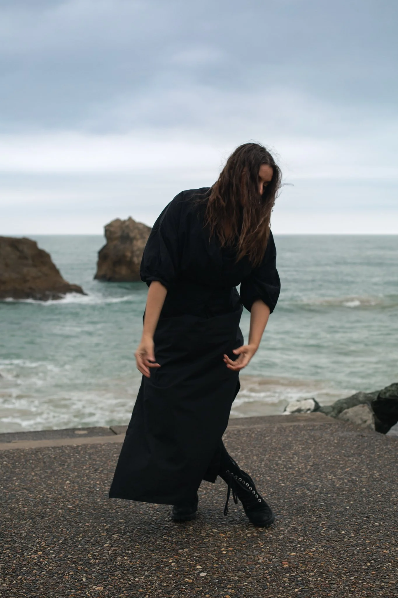 A woman in black clothing and boots standing on a pebbled surface near the ocean, with rocky formations and a cloudy sky in the background.