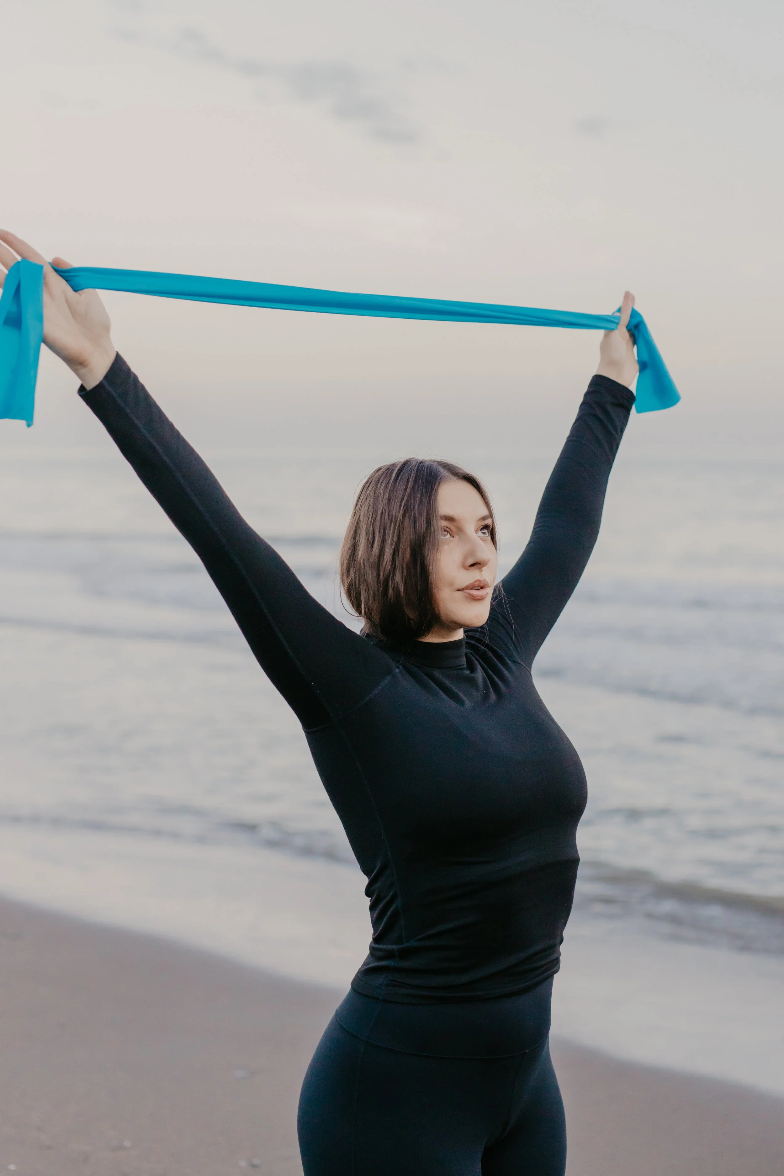 Woman in black shirt and leggings holding a blue resistance band above her head on a beach