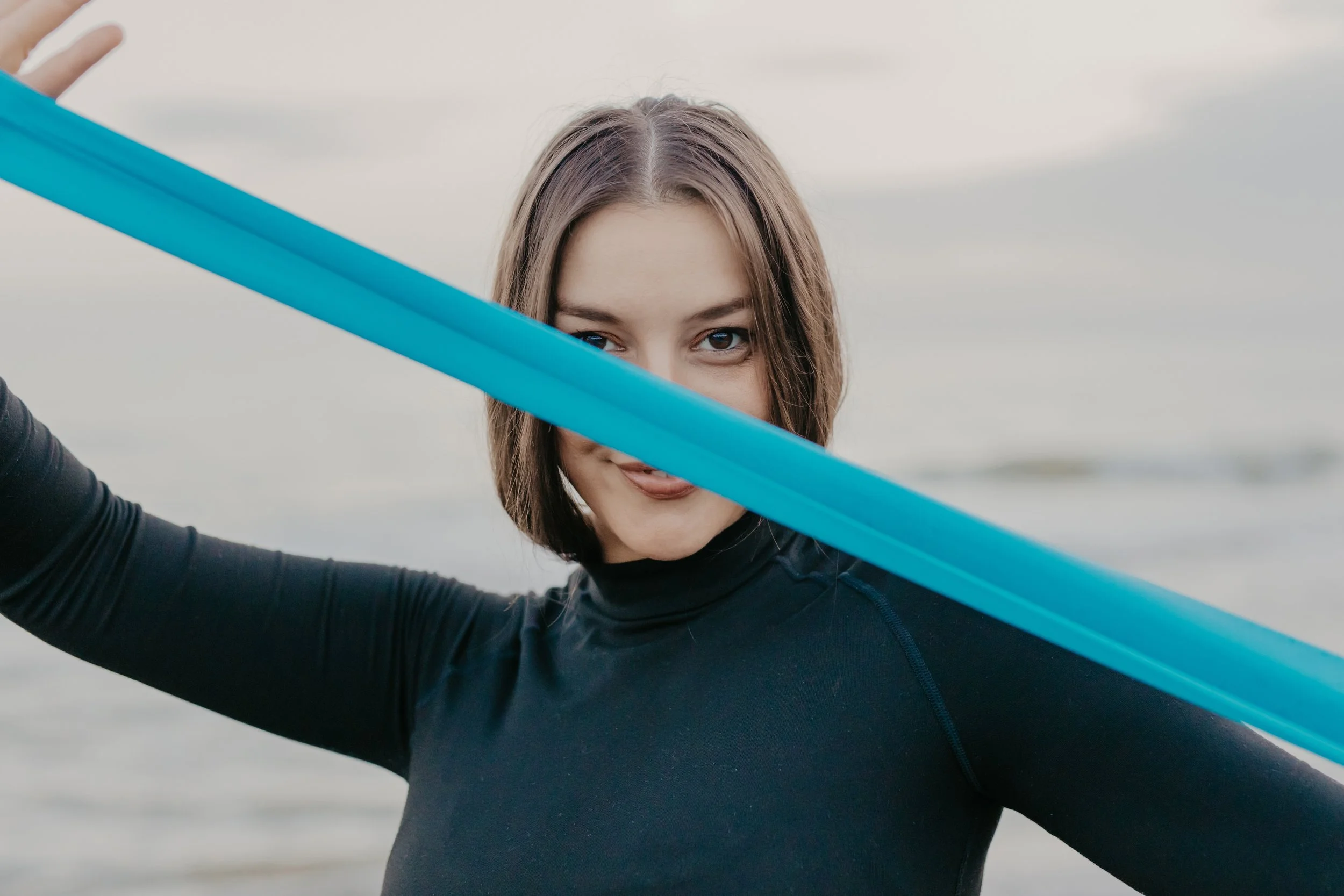 A young woman with brown hair and a mischievous smile, sticking out her tongue, holding two blue paddles in front of her face at the beach.