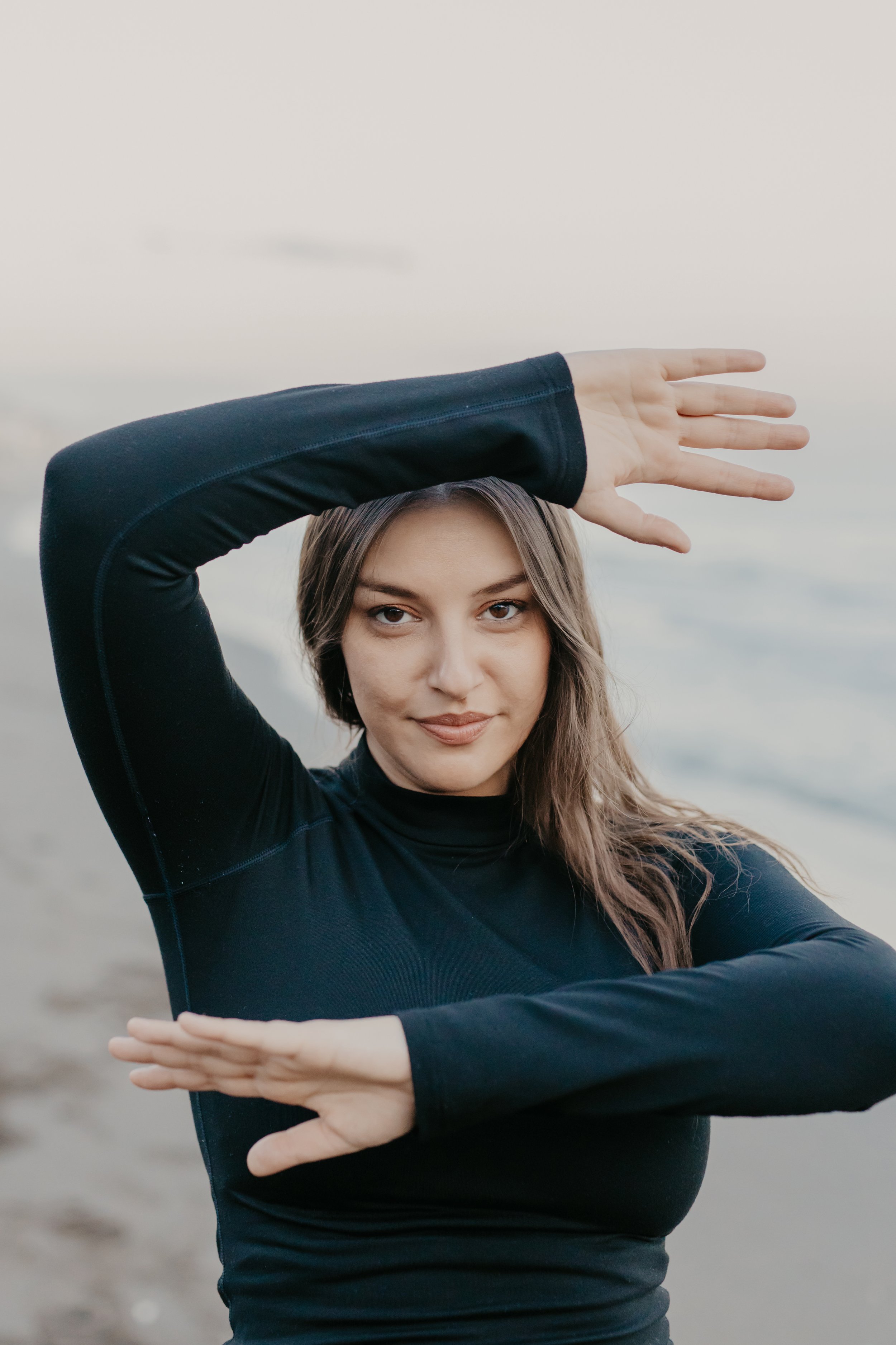 A young woman in a black long-sleeve top standing at the beach, with her left arm raised above her head and her right arm across her chest, making a frame shape with her arms and looking at the camera.