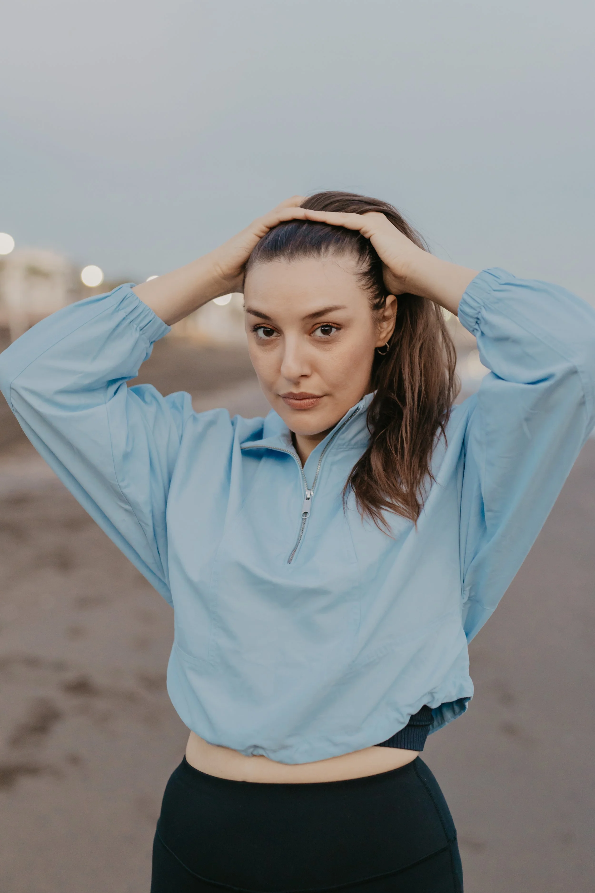 A young woman in a light blue windbreaker and black leggings standing outdoors on a cloudy day, holding her hair back with both hands, looking at the camera.