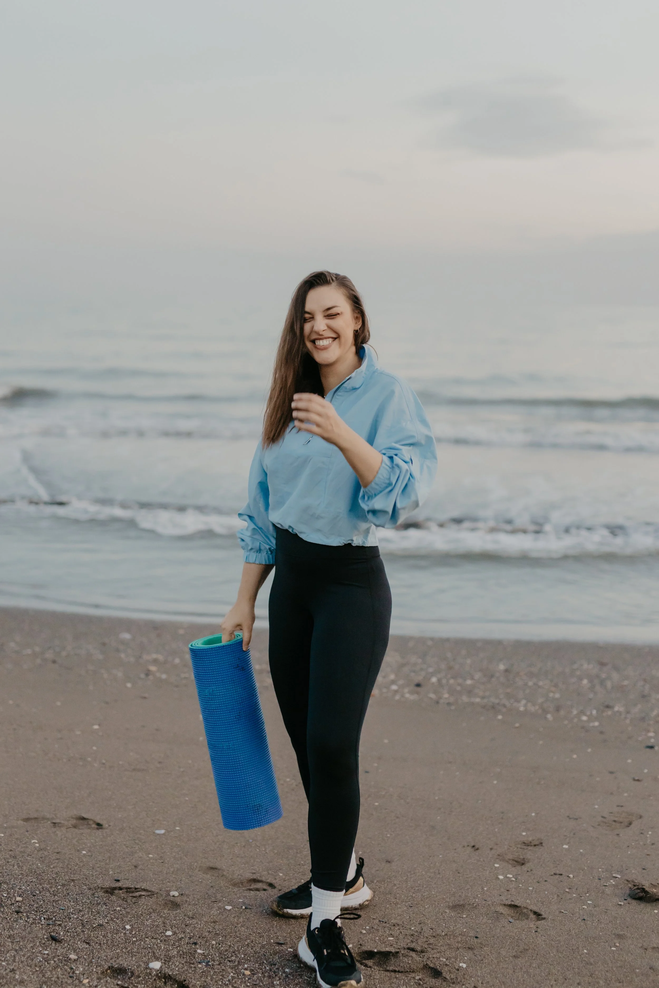 A woman standing on the beach holding a rolled-up yoga mat, wearing a light blue jacket, black leggings, and black sneakers, smiling.