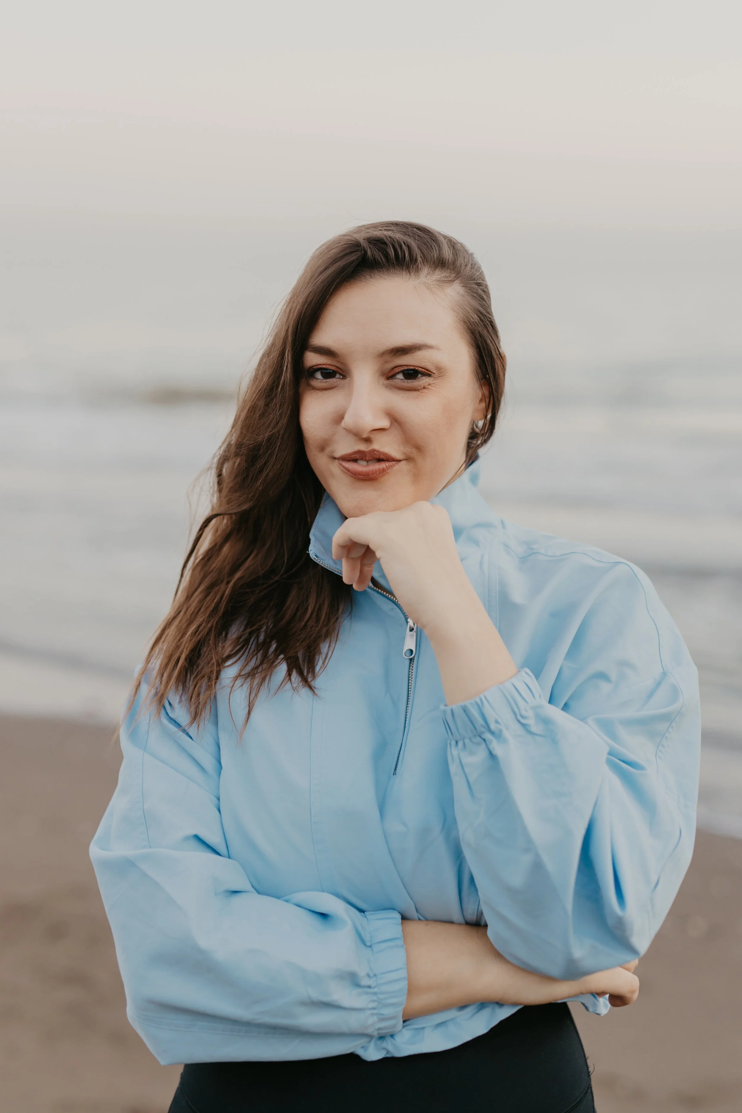 A young woman with long brown hair, wearing a light blue jacket, stands on a beach during sunset, looking at the camera with a slight smile.