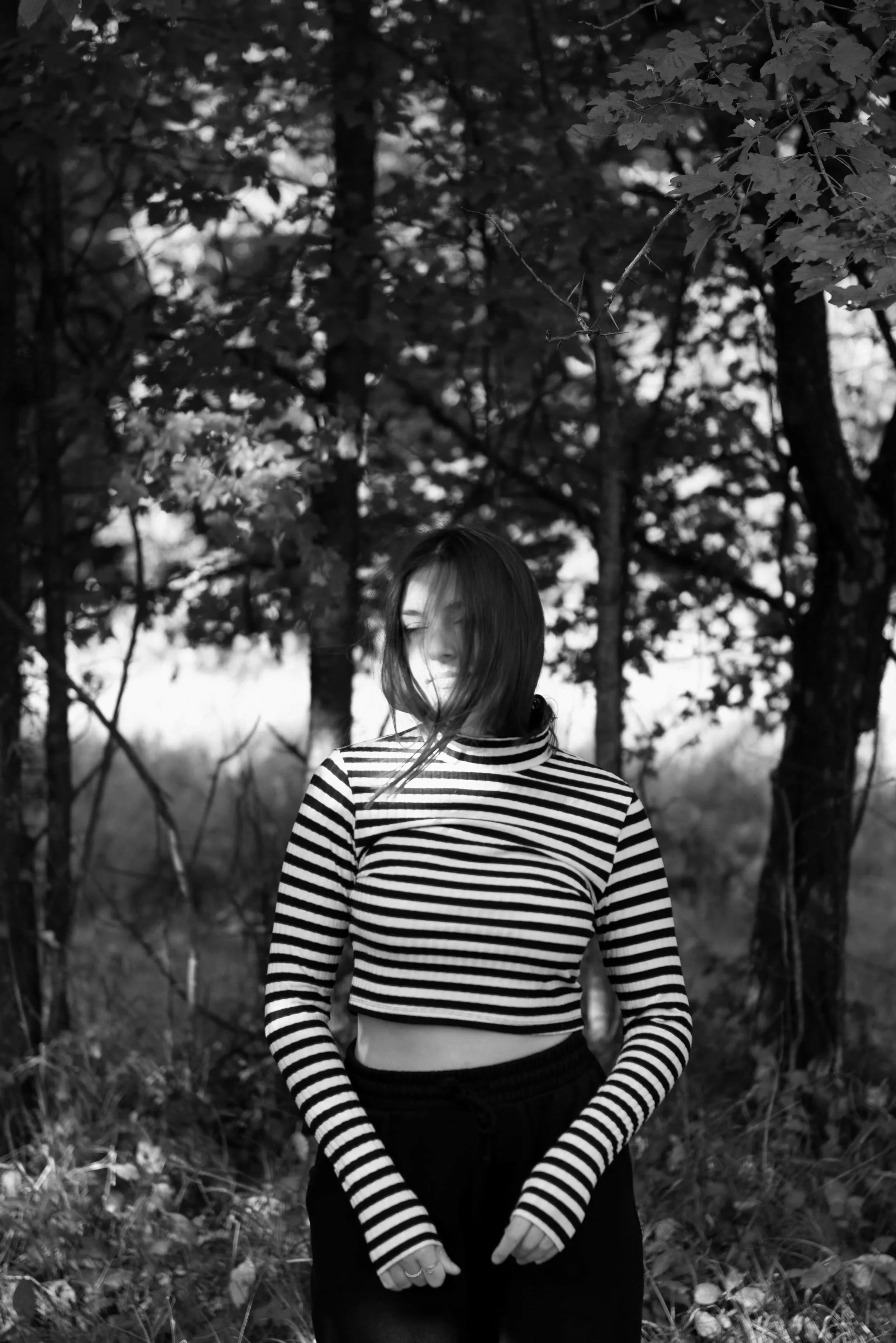 A black and white photo of a woman with shoulder-length hair standing outdoors among trees, wearing a striped long-sleeve crop top and black pants, with her eyes closed and hair blowing across her face.