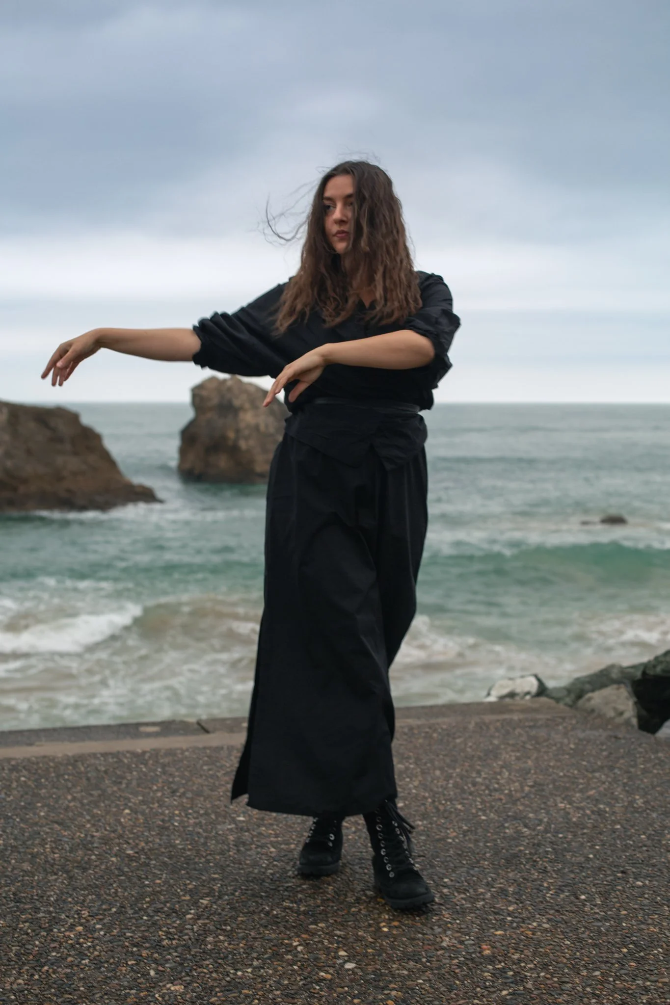 A woman with long curly hair standing on a rocky shore with the ocean and large rock formations in the background, wearing a black outfit and black boots.