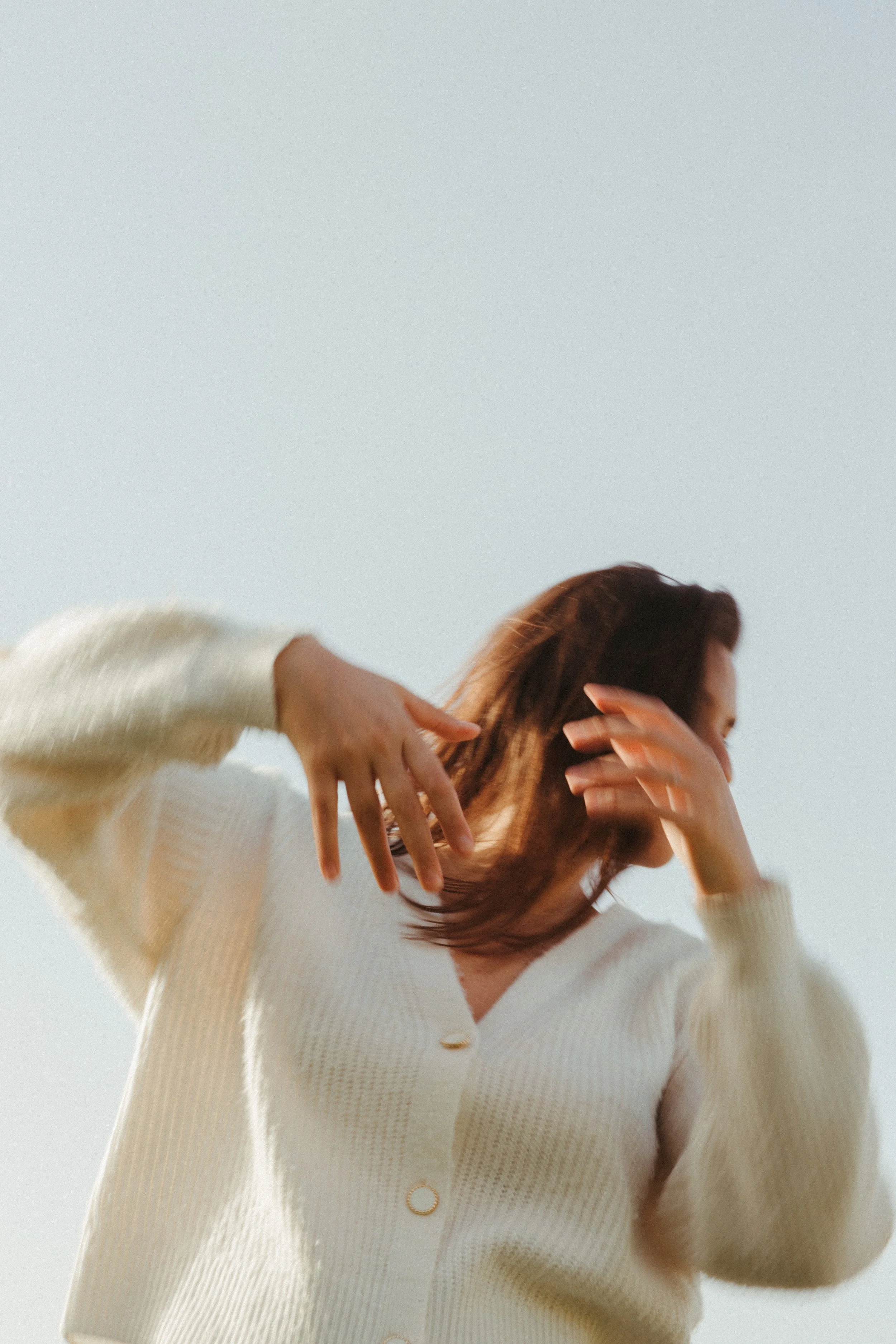 A woman with brown hair wearing a cream-colored cardigan, touching her hair and face, against a clear sky.